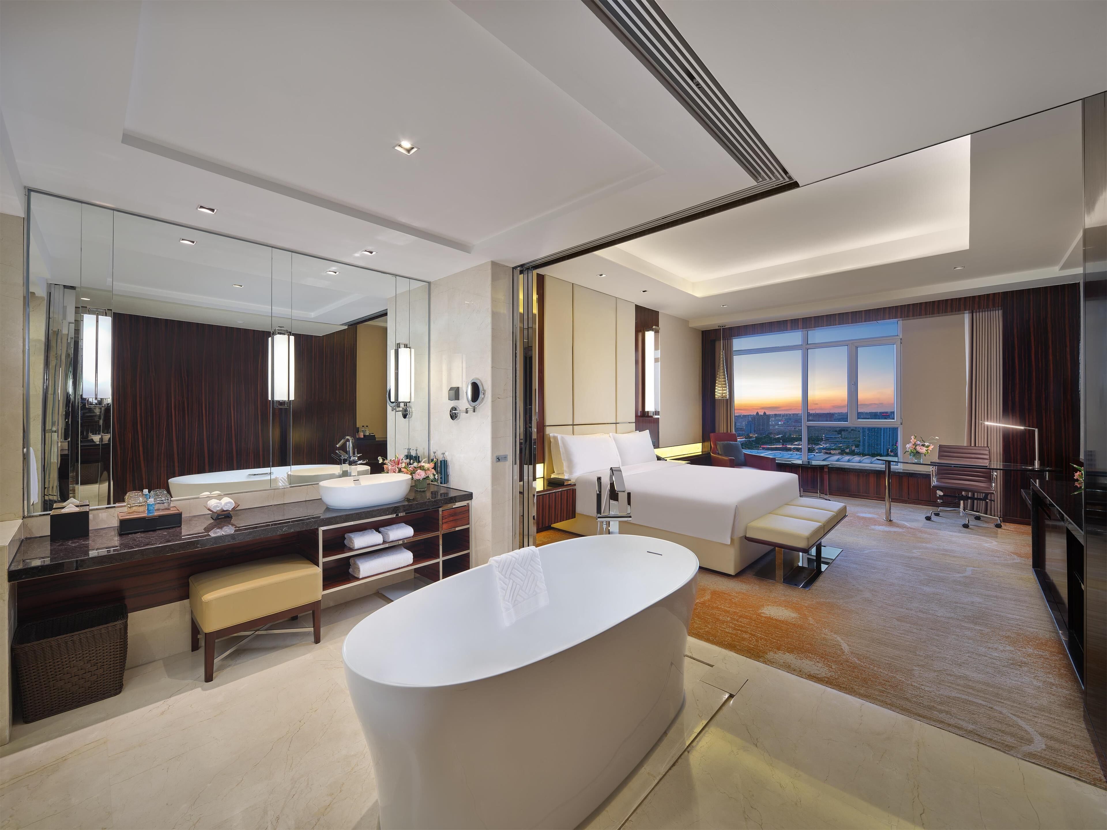 Una habitación de hotel moderna con una bañera grande, una cama doble, un escritorio y un amplio ventanal con vista a la ciudad.