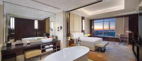A modern hotel room with a large bathtub, a double bed, a desk, and a large window offering a city view.