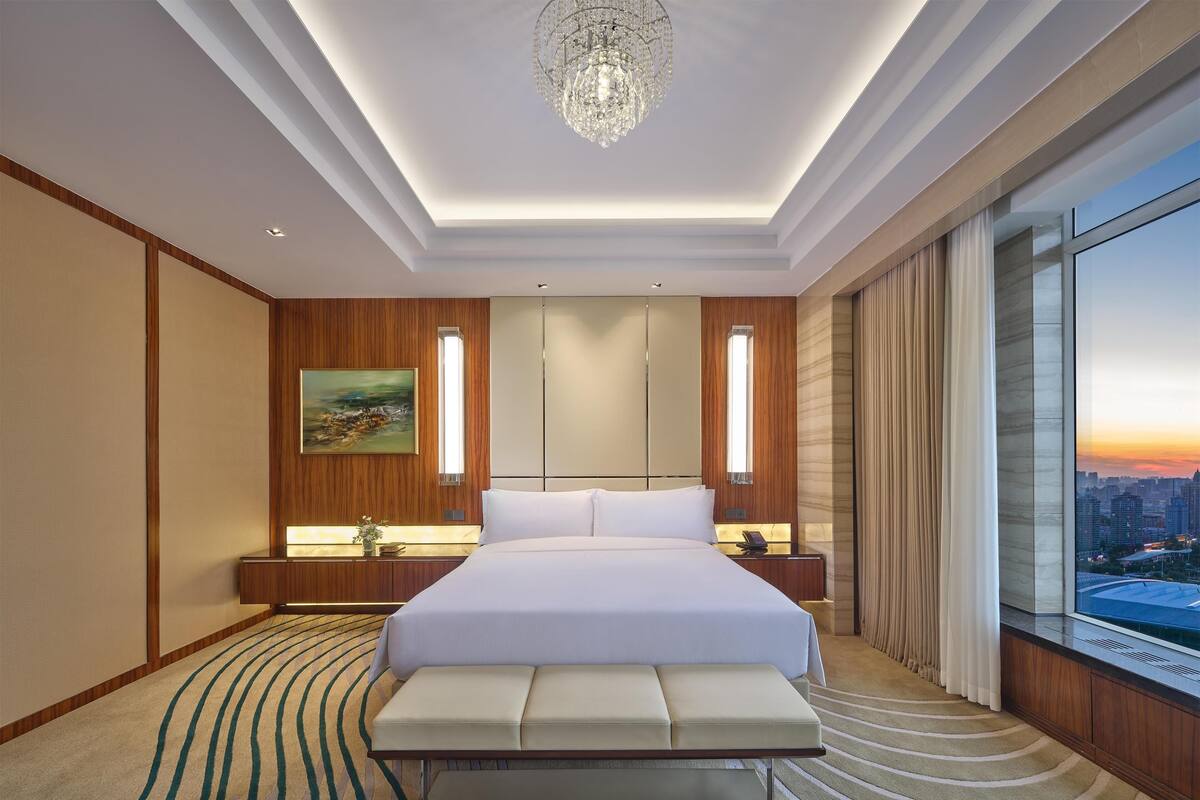 Ein modernes Hotelzimmer mit einem großen Bett, einer Bank, einem Gemälde und Blick auf die Stadt.