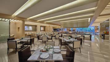 A spacious restaurant with multiple dining tables, a central bar area, and large windows offering a view of the outdoors.