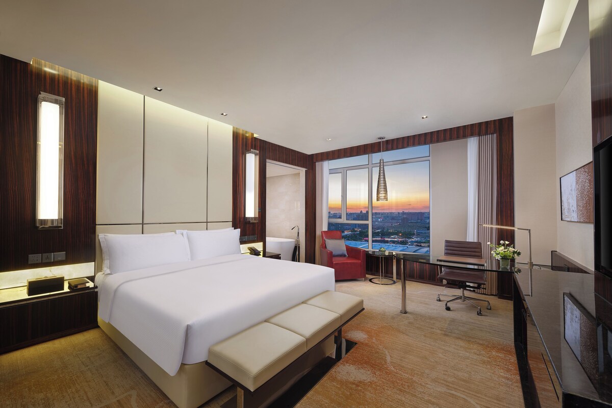 A modern hotel room with a large bed, a desk, a chair, and a view of the cityscape at sunset.