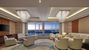 A spacious living room with a large TV, comfortable seating, a view of the city, and elegant chandeliers. - Hilton Harbin Exhibition Center (Harbin)