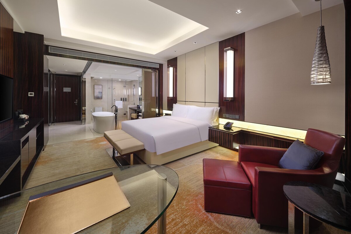 A modern hotel room with a large bed, a glass coffee table, a red armchair, a desk, and a TV.