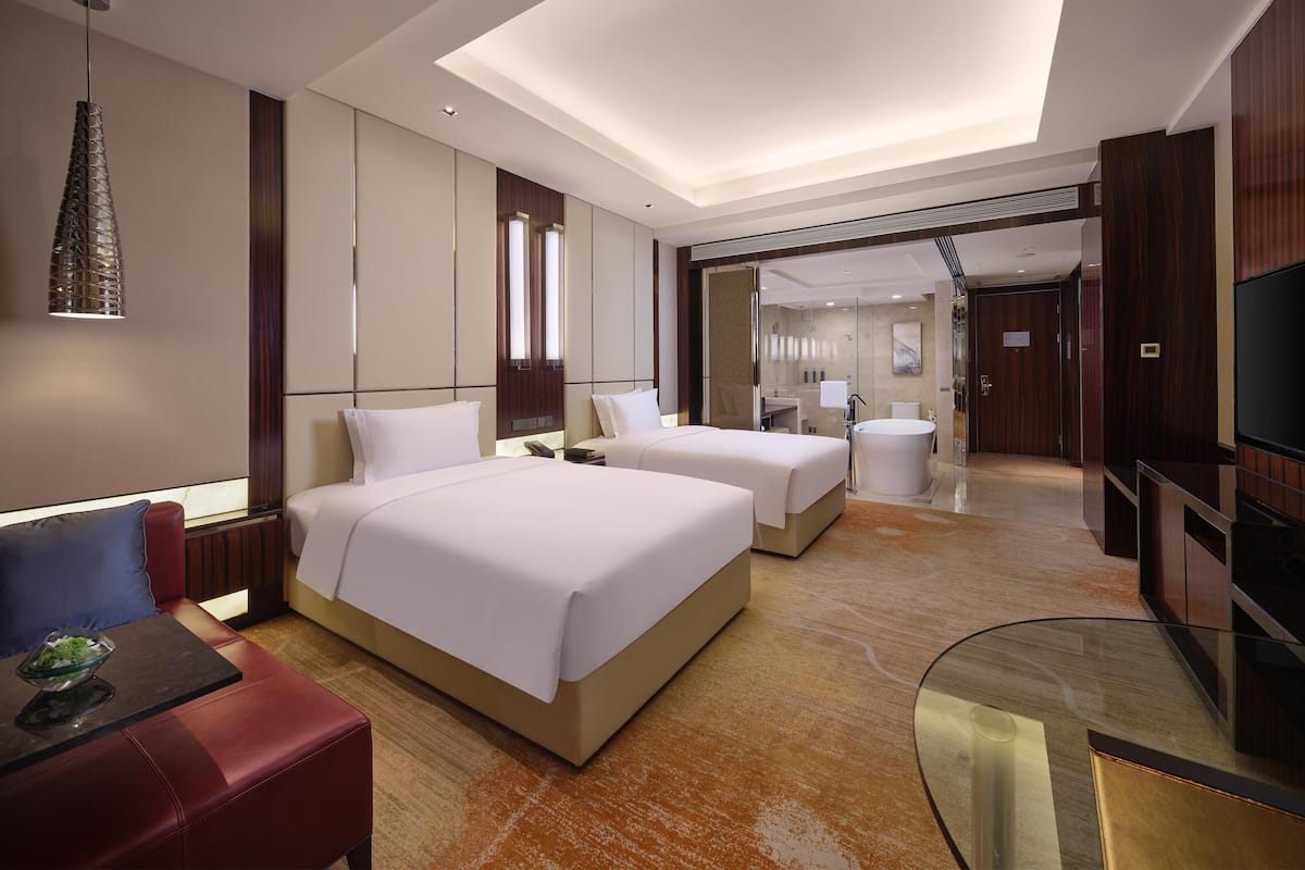 Ein modernes Hotelzimmer mit zwei Betten, einer Sitzecke, einem Fernseher und einem durchgegangene Tür erkennbaren Badezimmer.