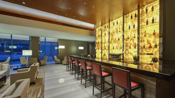 Zona de bar moderna con una barra larga, taburetes rojos y una vitrina con botellas de licores.