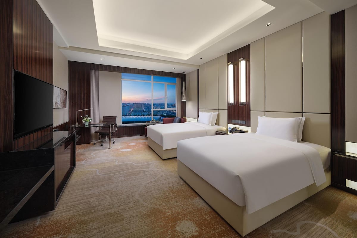 Ein Hotelzimmer mit zwei Betten, einem Schreibtisch, einem Fernseher und einem großen Fenster mit Blick auf die Stadt.