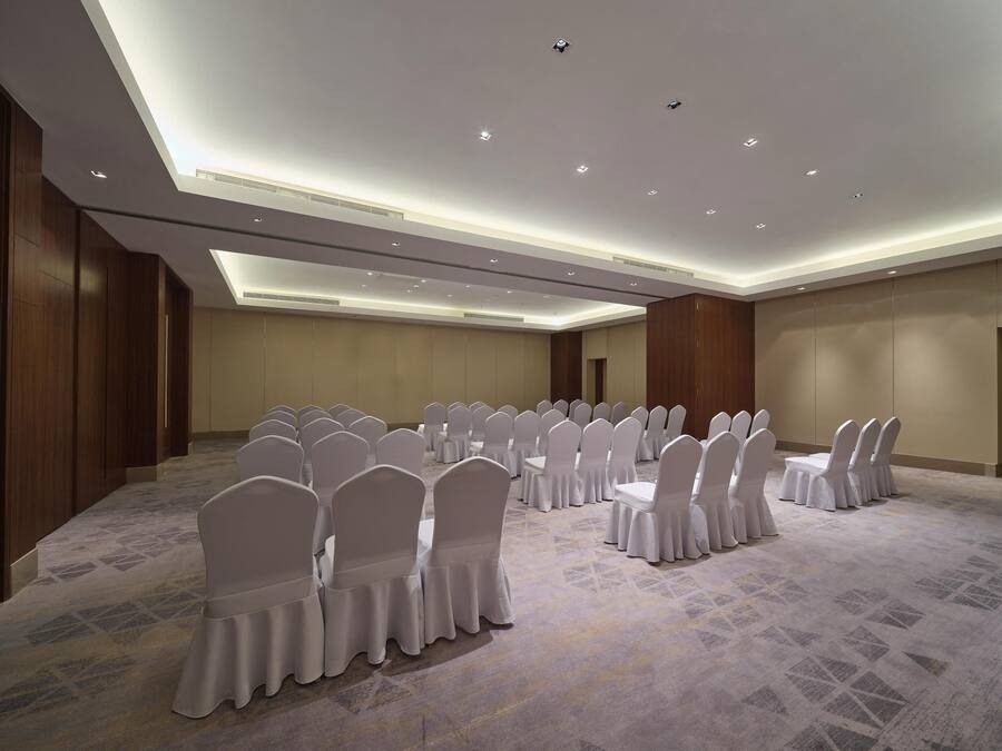 A conference room with rows of white chairs and tables, a patterned carpet, and recessed ceiling lighting.