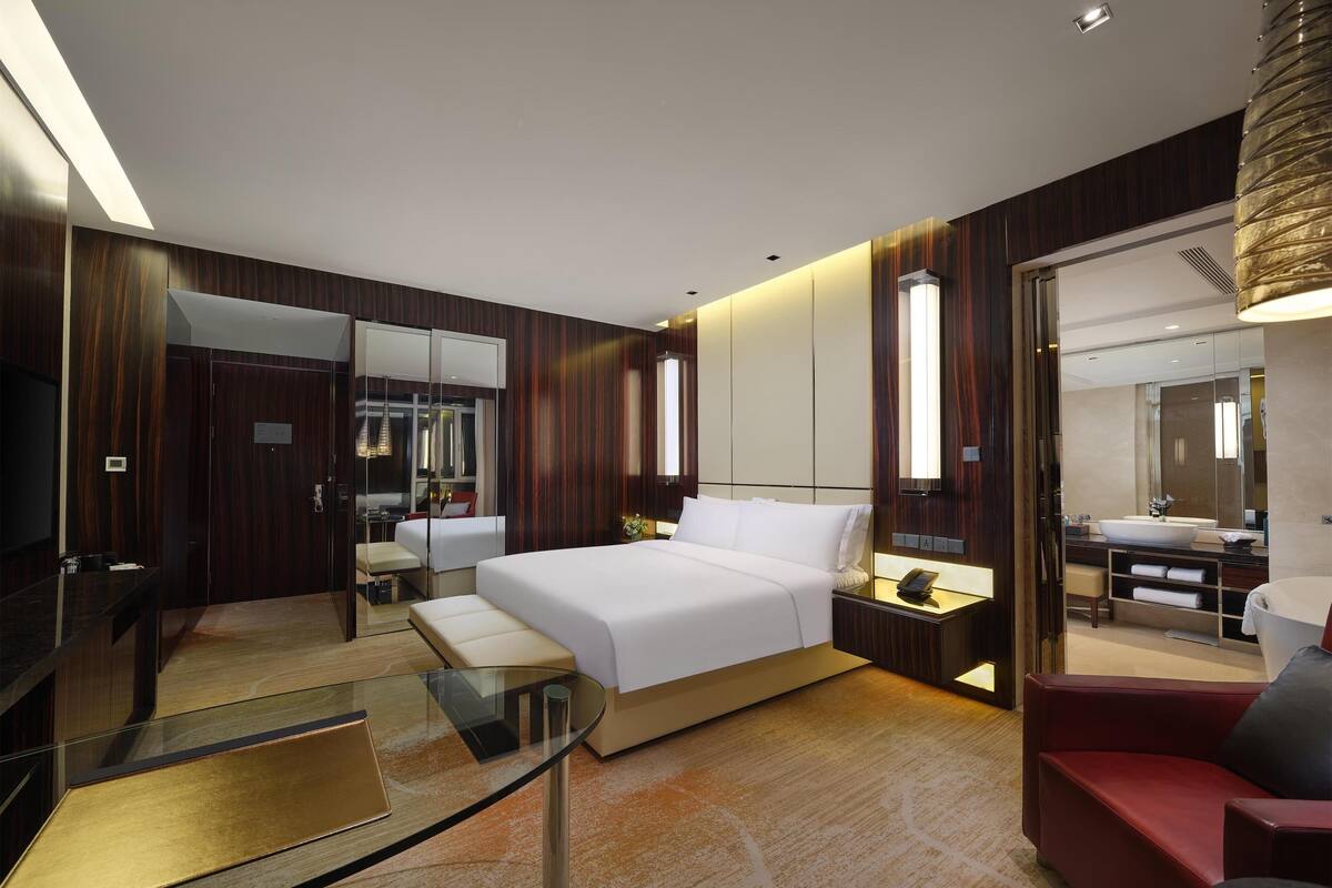 Ein modernes Hotelzimmer mit einem großen Bett, einem Glastisch, einem roten Sofa und einem Badezimmer mit einem großen Spiegel und Waschbecken.