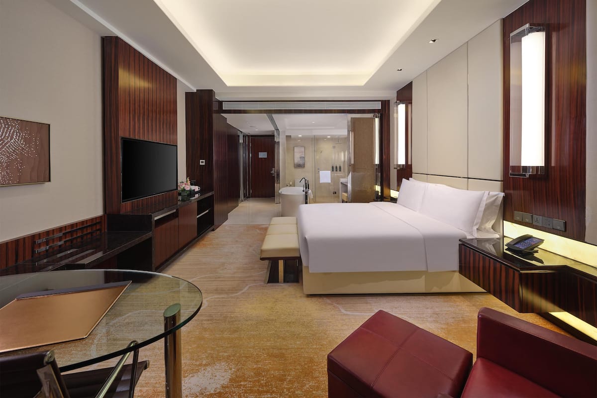 Ein modernes Hotelzimmer mit einem großen Bett, einem Schreibtisch mit Glasplatte, einem Flachbildfernseher und einer Sitzecke mit einem roten Sessel.