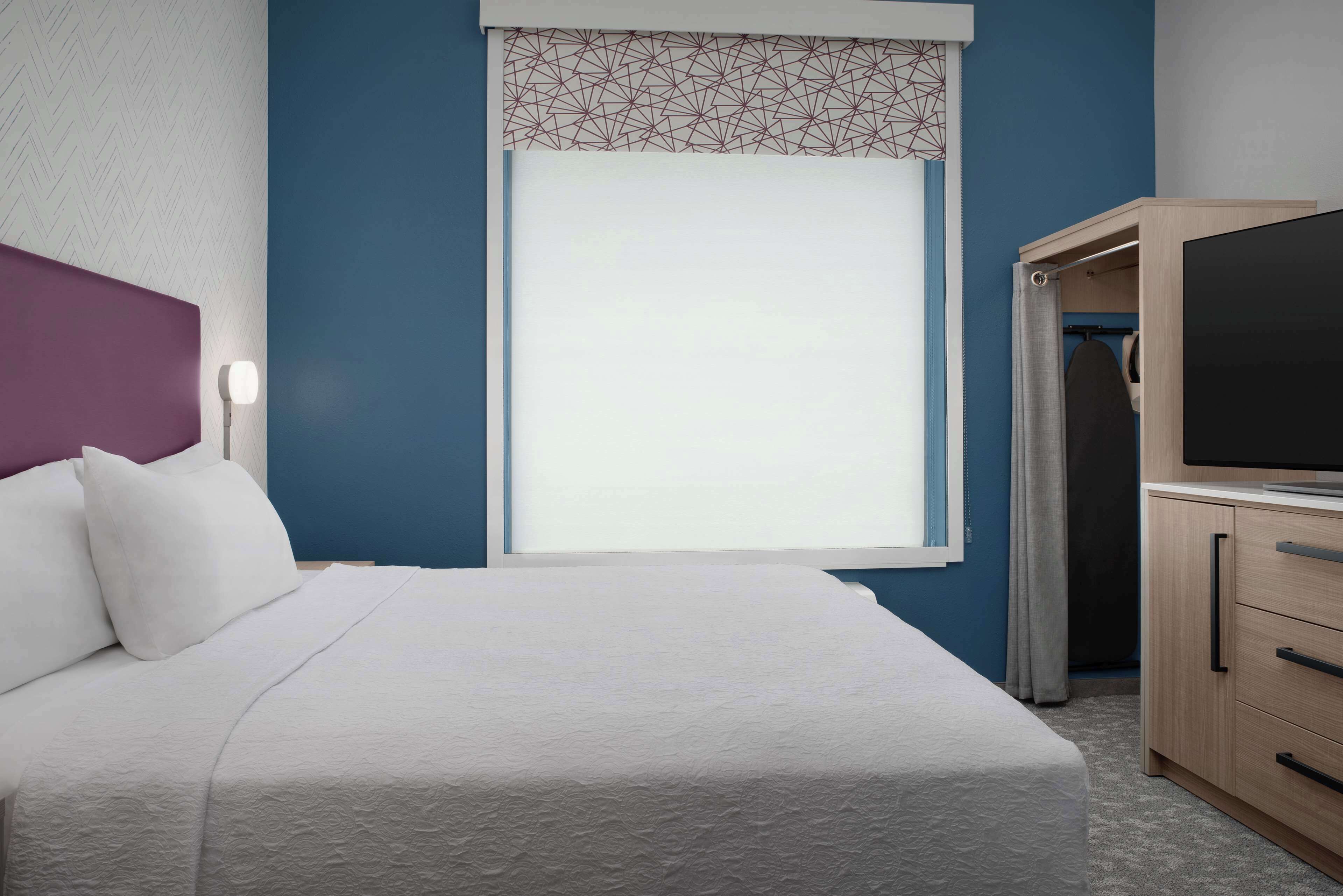 A neatly made bed with white bedding, a headboard, and a window with a geometric patterned blind.