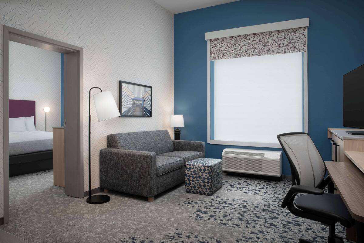 A hotel room with a bed, a grey sofa, a small ottoman, a desk, a chair, a standing lamp, and a large window with a geometric pattern.