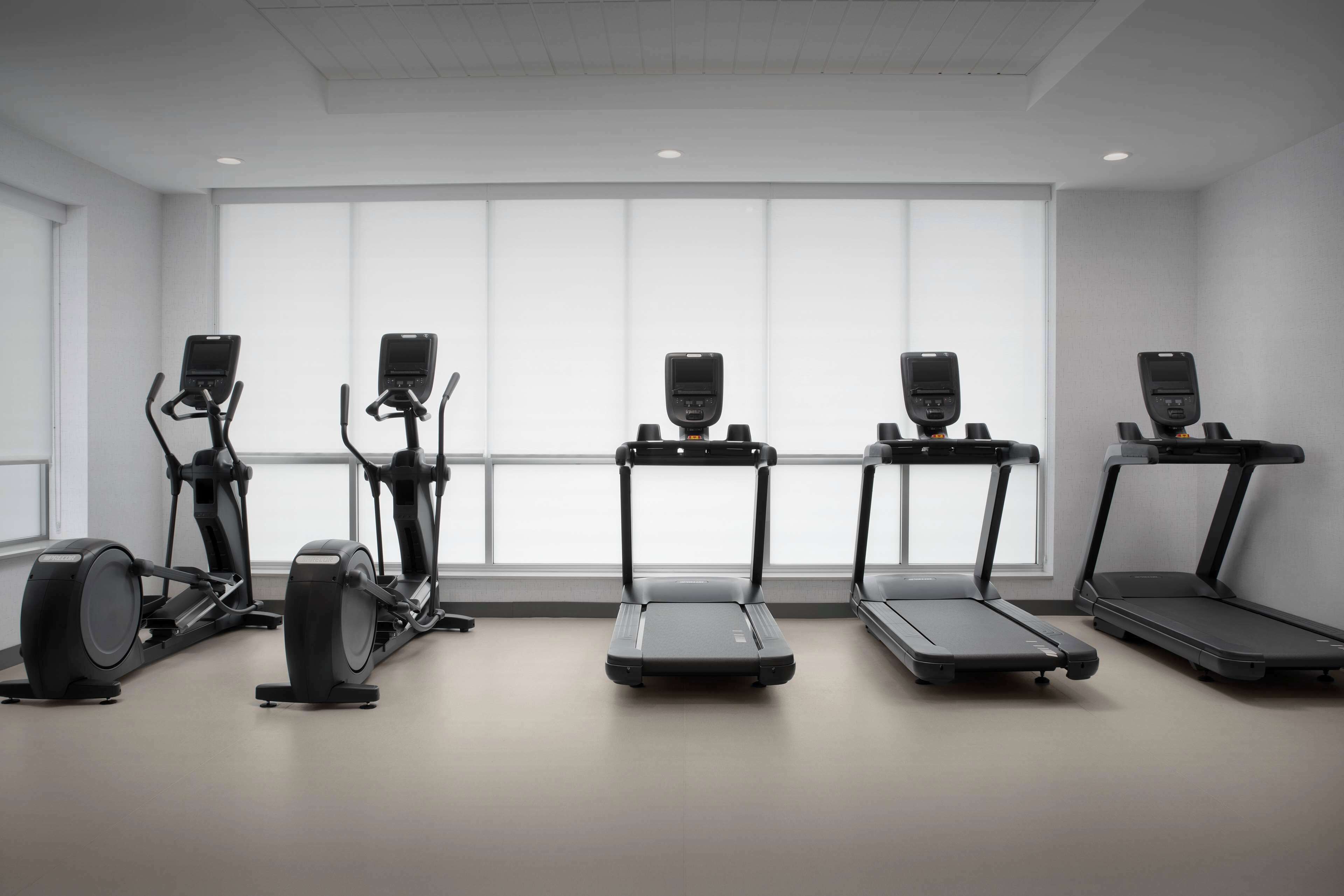 Une salle de sport équipée de cinq appareils d’exercice, dont des tapis de course et un vélo elliptique, dans une pièce dotée de grandes fenêtres et d’un plafond blanc.