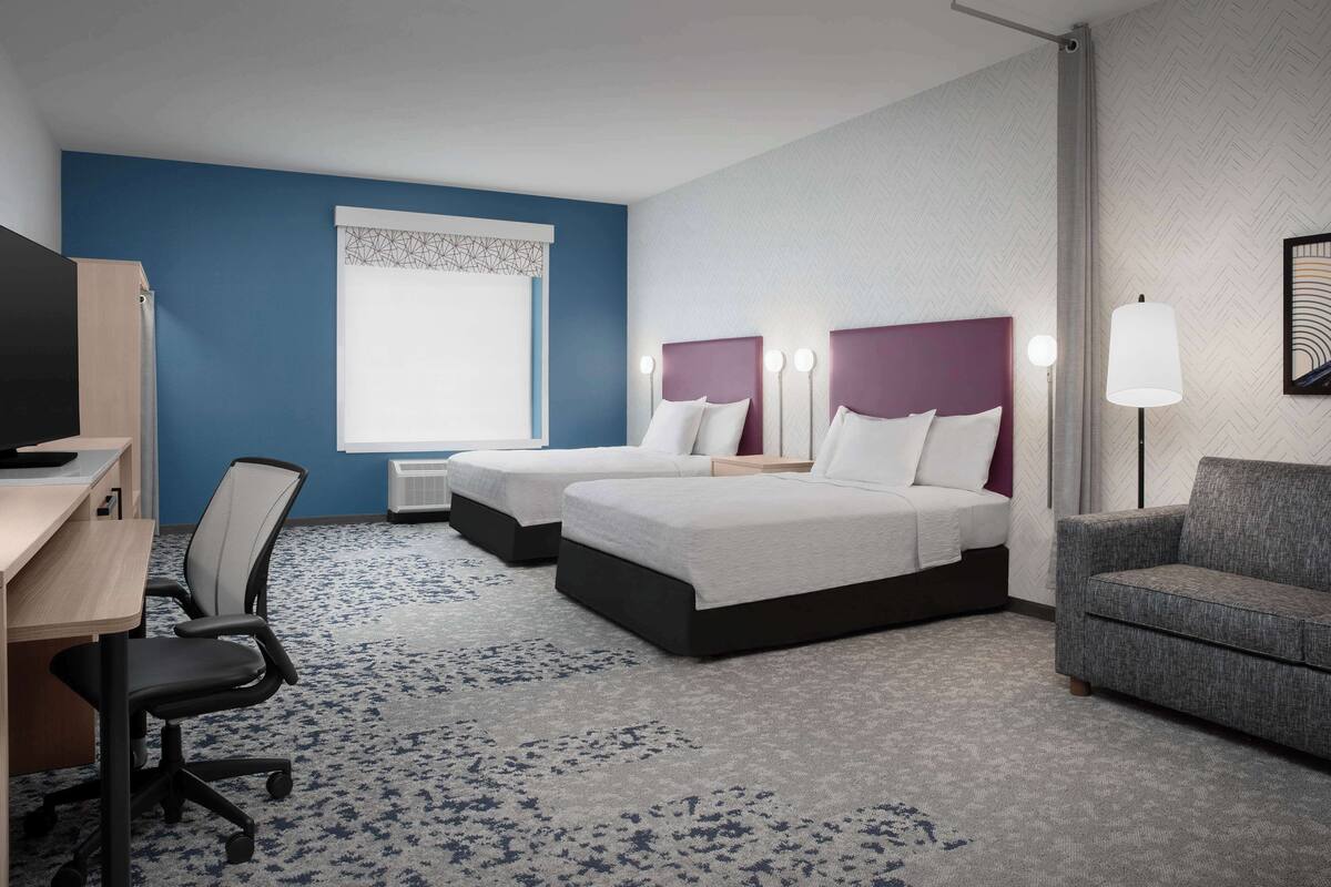 A hotel room with two beds, a desk, a chair, a sofa, and a window with a geometric curtain.