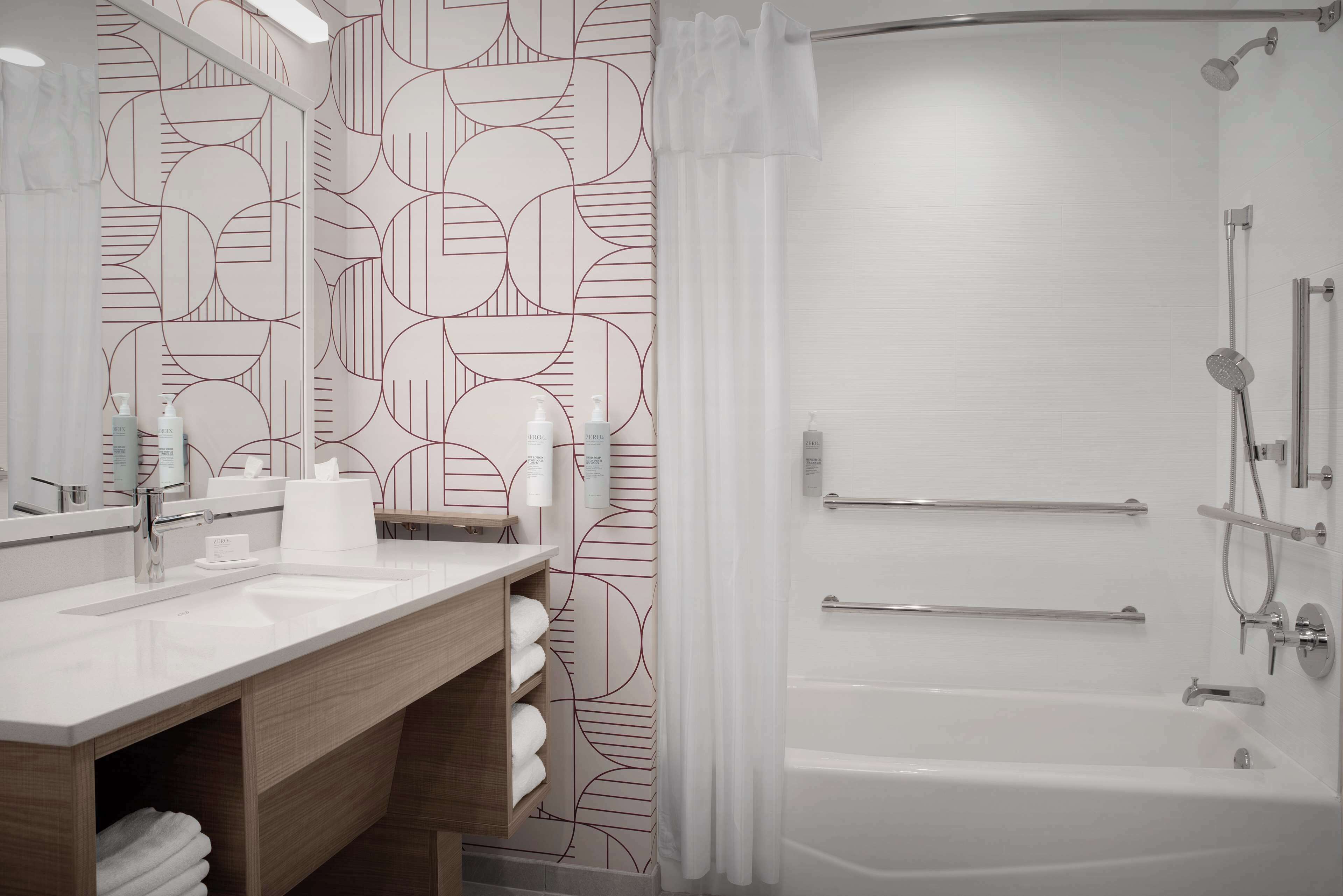 A modern bathroom with a geometric patterned wall, a white sink with a mirror, and a walk-in shower with grab bars.