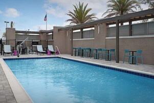 Una piscina con baldosas azules, rodeada de sillas turquesa y sillones morados, con un muro de ladrillo y una bandera estadounidense al fondo.
