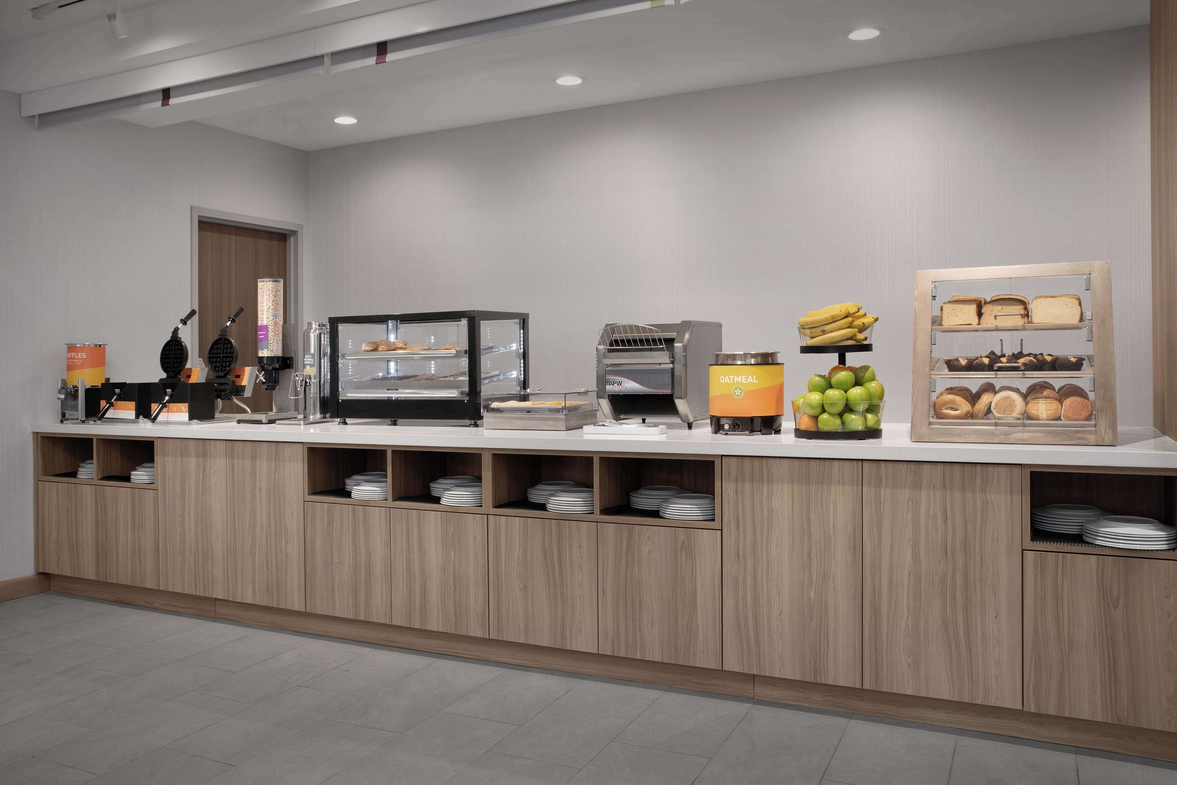 Un bar à petit-déjeuner moderne proposant une variété d’aliments, notamment des gaufres, du porridge, des bananes, des pommes et des viennoiseries.