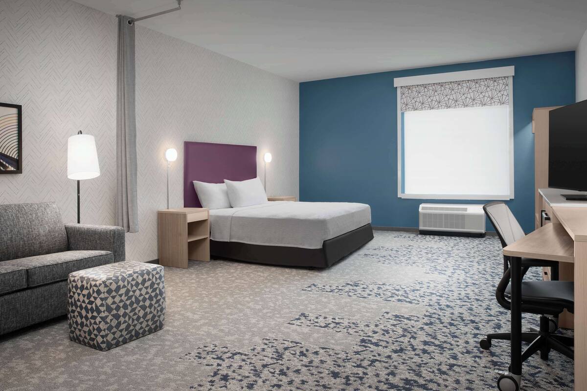 A hotel room with a bed, a desk, a chair, a sofa, and a patterned ottoman.