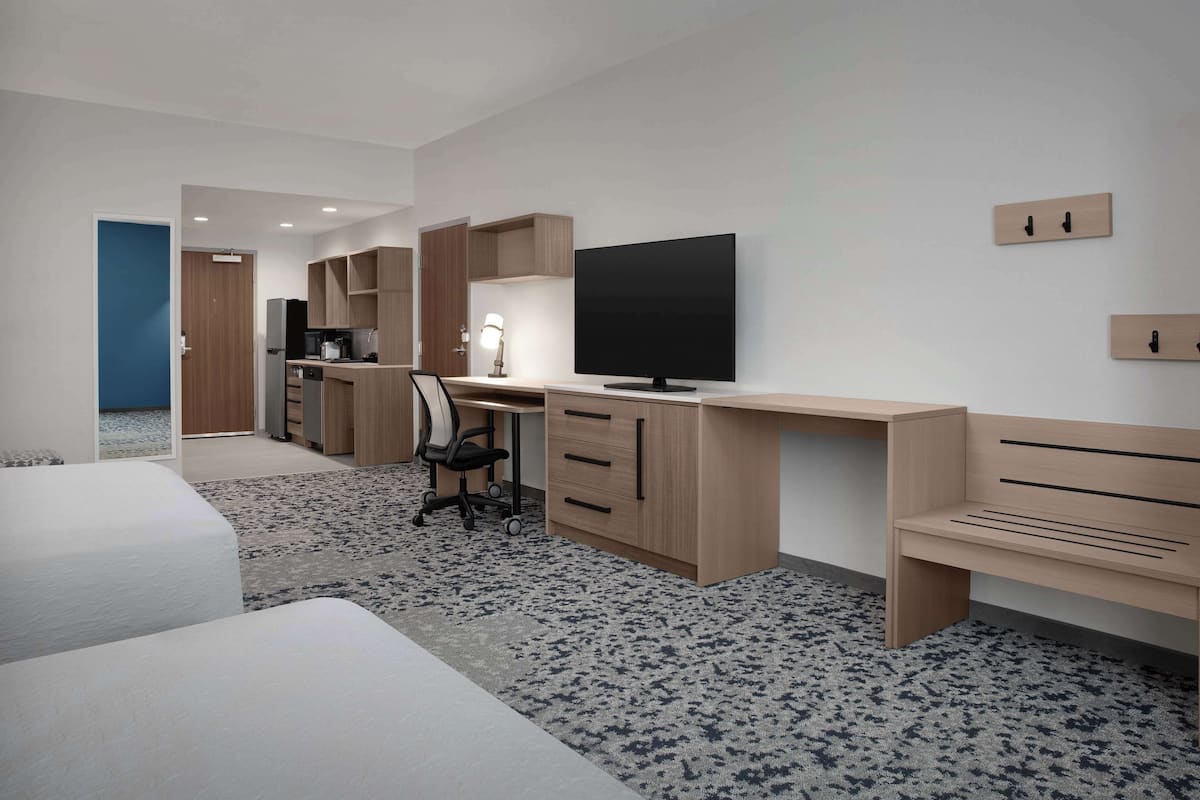 A modern hotel room with a large bed, a desk with a computer, a TV, and a kitchenette.
