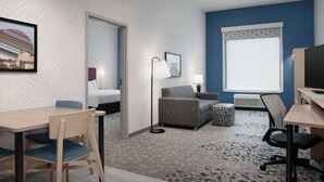Habitación de hotel con pared de diseño espiga, un sofá gris, una pequeña mesa con sillas, un escritorio con una silla y un ventanal con un motivo geométrico.