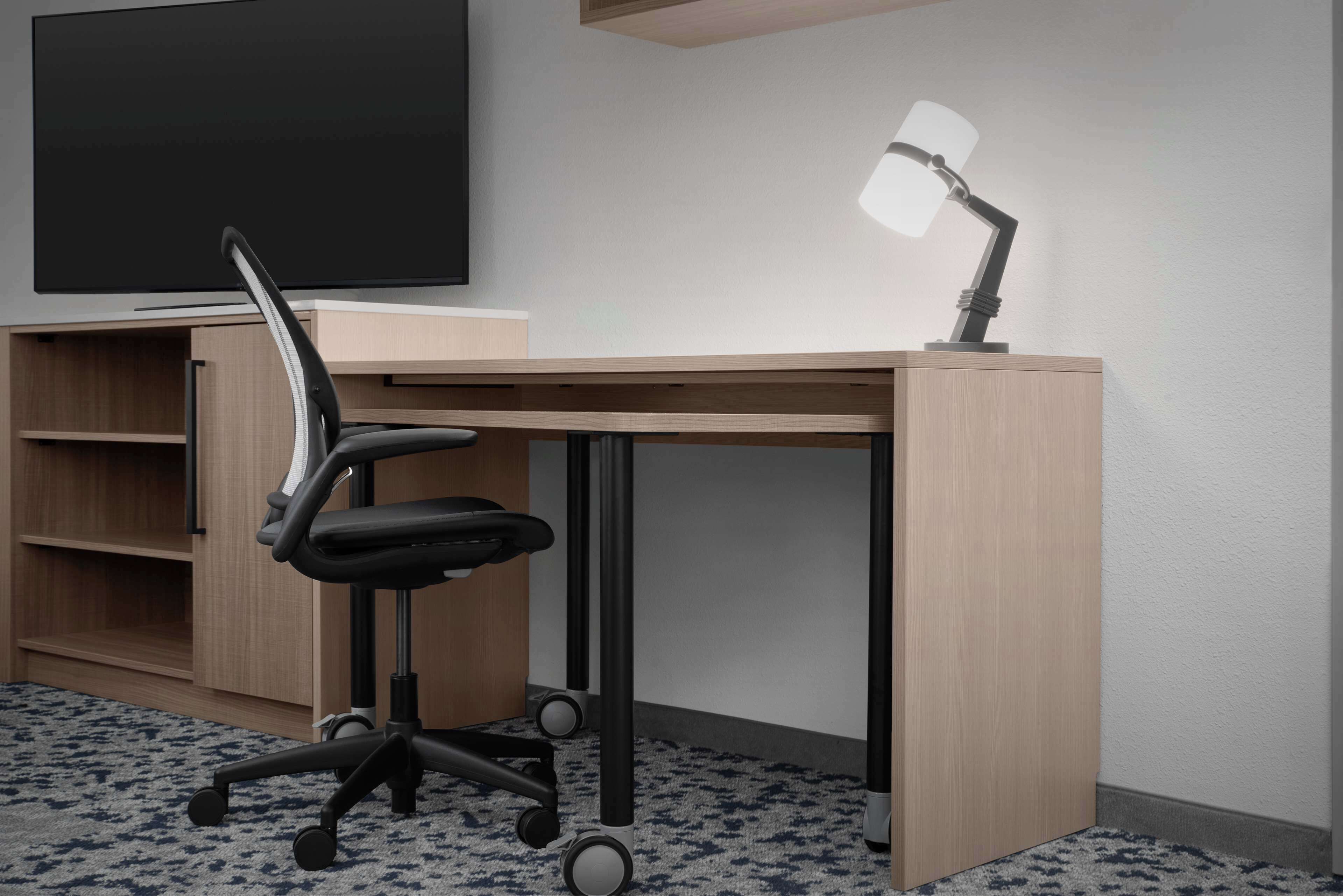 A modern office desk with a black office chair, a lamp, and a flat-screen TV on a wooden shelf.