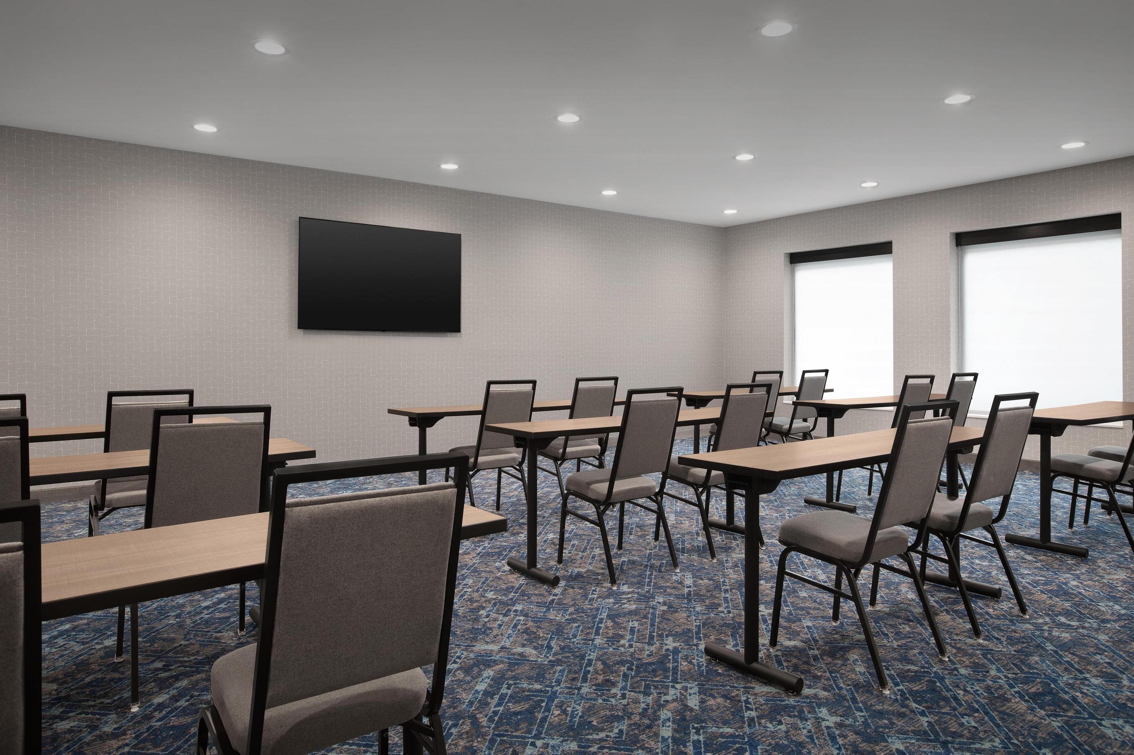 Un salón de conferencias con mesas, sillas, una pantalla grande y ventanas.