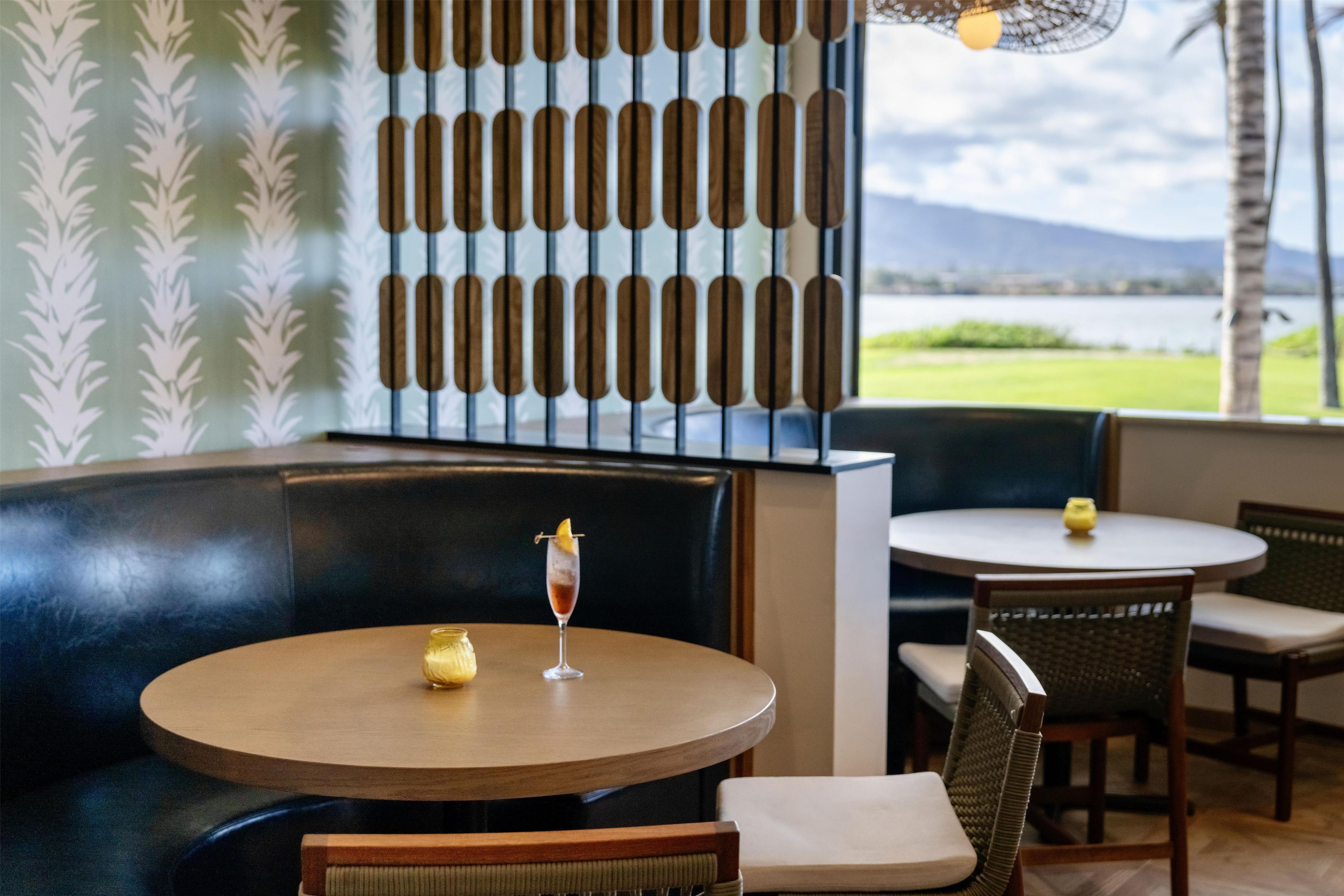 Ein Restaurant mit runden Tischen und Sitzecken, eine Wand mit Blattmuster-Dekoration und Blick auf ein Gewässer.