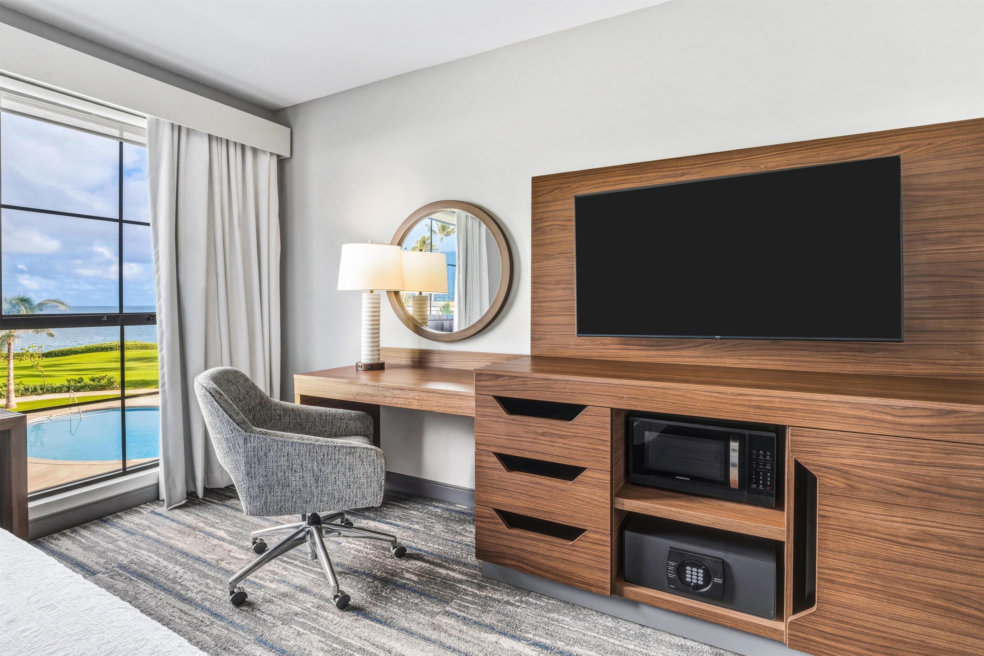 Ein Hotelzimmer mit großem Fenster, Schreibtisch mit Flachbildfernseher, Stuhl, Lampe und Mikrowelle.