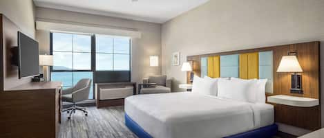 A hotel room with a large bed, a desk, a chair, a TV, and a view of the sea.