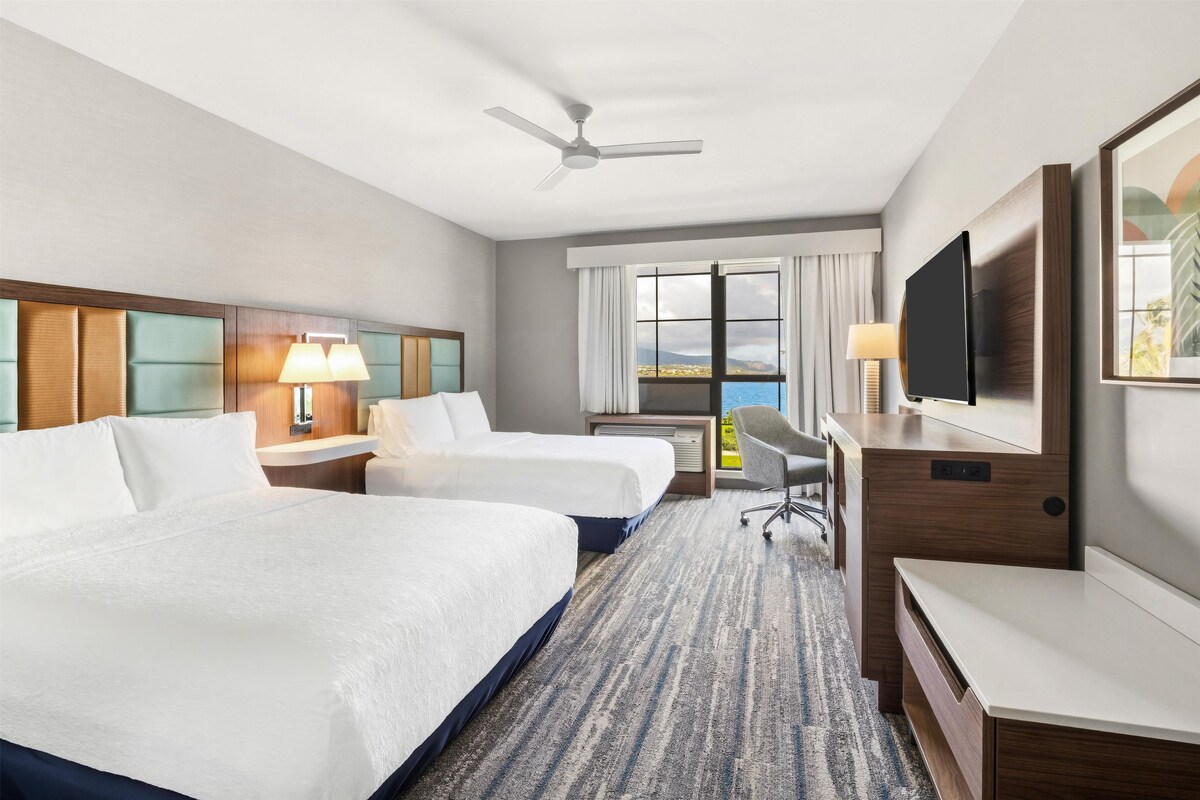 A hotel room with two beds, a desk, a TV, and a view of the outdoors.