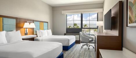 A hotel room with two beds, a desk, a chair, a TV, and a window with a view of palm trees.
