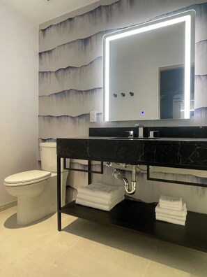 Un baño con un lavamanos de mármol negro, un inodoro blanco y un espejo con luz integrado en la pared.