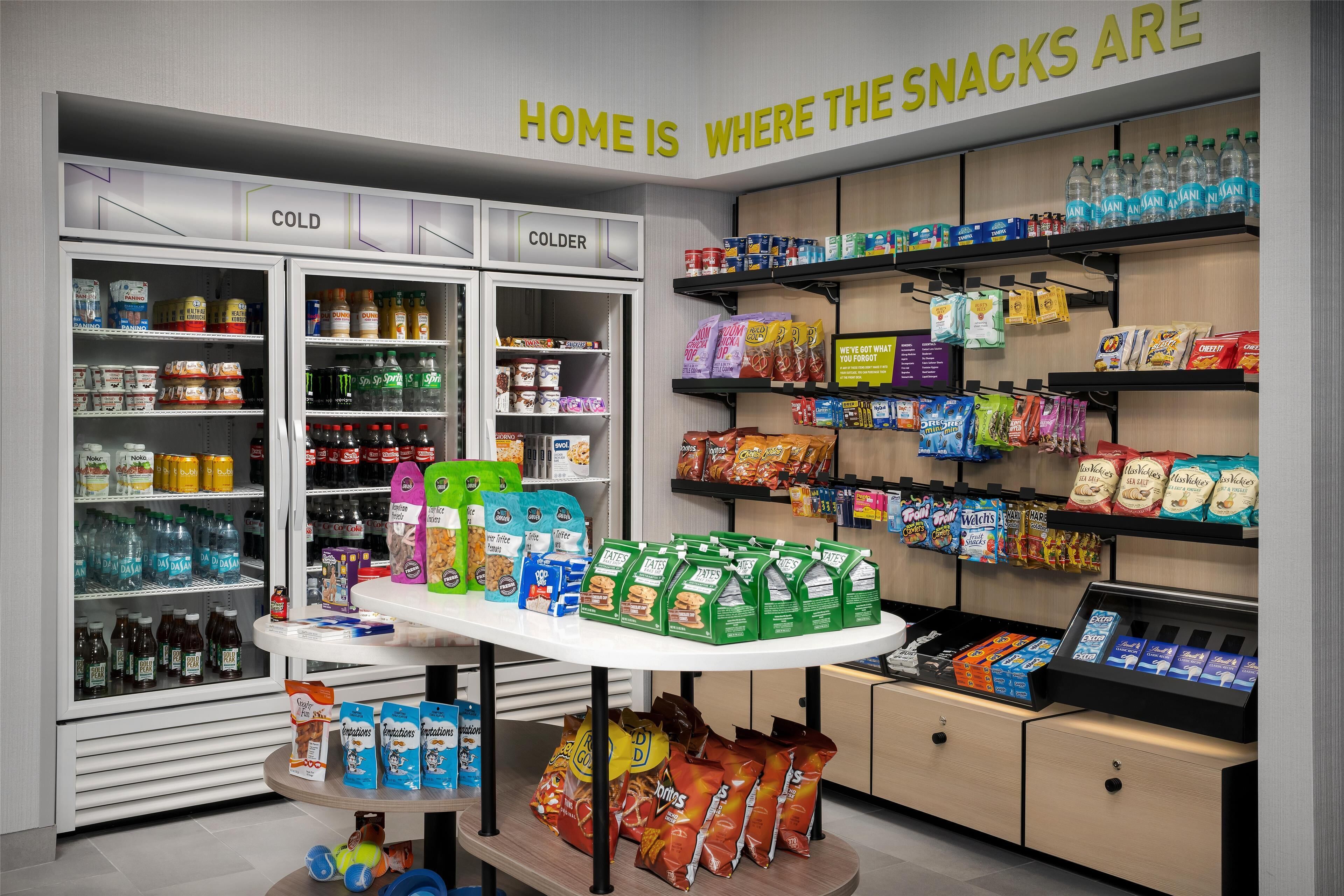 Una sección de snacks con estantes refrigerados y no refrigerados, una variedad de productos envasados, y un letrero que dice “HOME IS WHERE THE SNACKS ARE”.