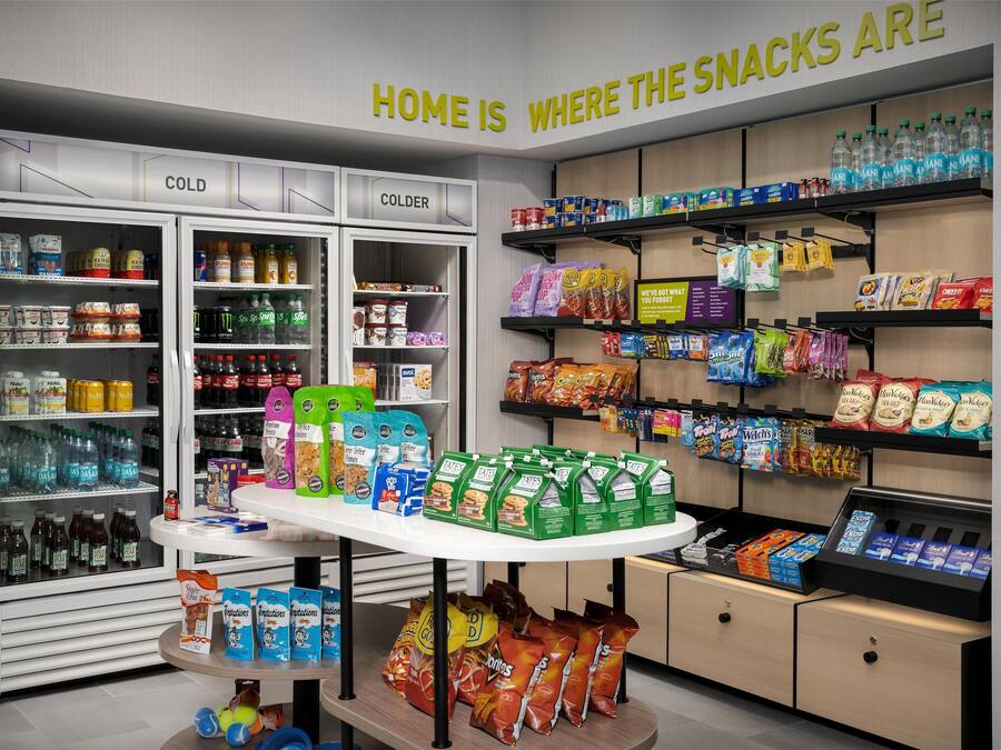 Una sección de snacks con estantes refrigerados y no refrigerados, una variedad de productos envasados, y un letrero que dice “HOME IS WHERE THE SNACKS ARE”.