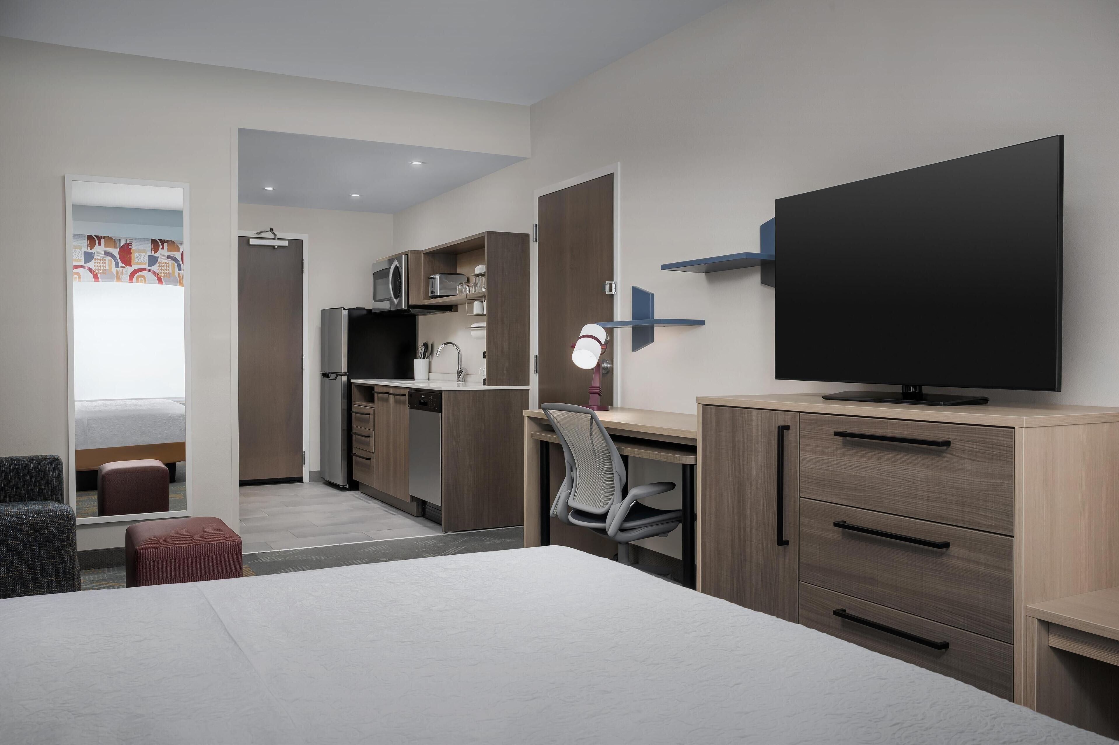 A modern hotel room with a large bed, a desk with a chair, a television, a microwave, and a kitchenette.