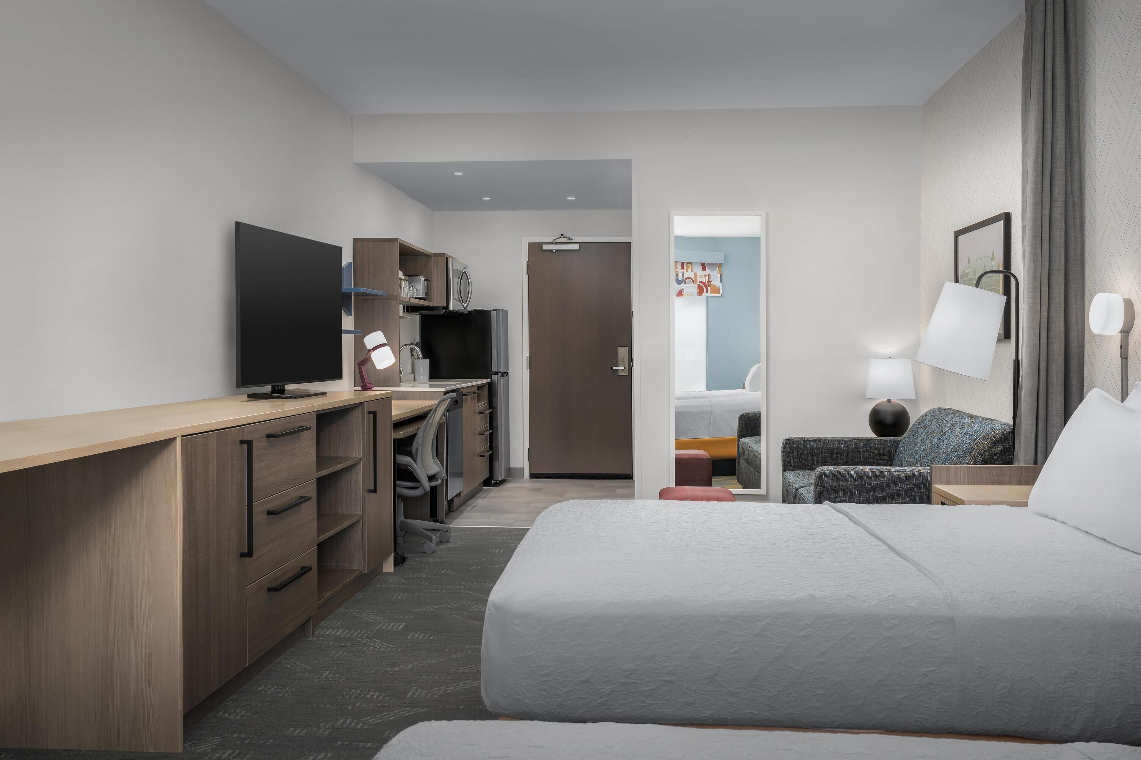 Ett modernt hotellrum med en stor säng, ett skrivbord med en platt-TV, en garderob, en sittgrupp med en stol och ett bord samt en dörr som leder till badrummet.