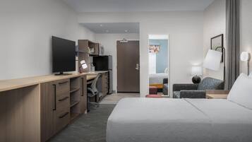 Ett modernt hotellrum med en stor säng, ett skrivbord med en platt-TV, en garderob, en sittgrupp med en stol och ett bord samt en dörr som leder till badrummet.