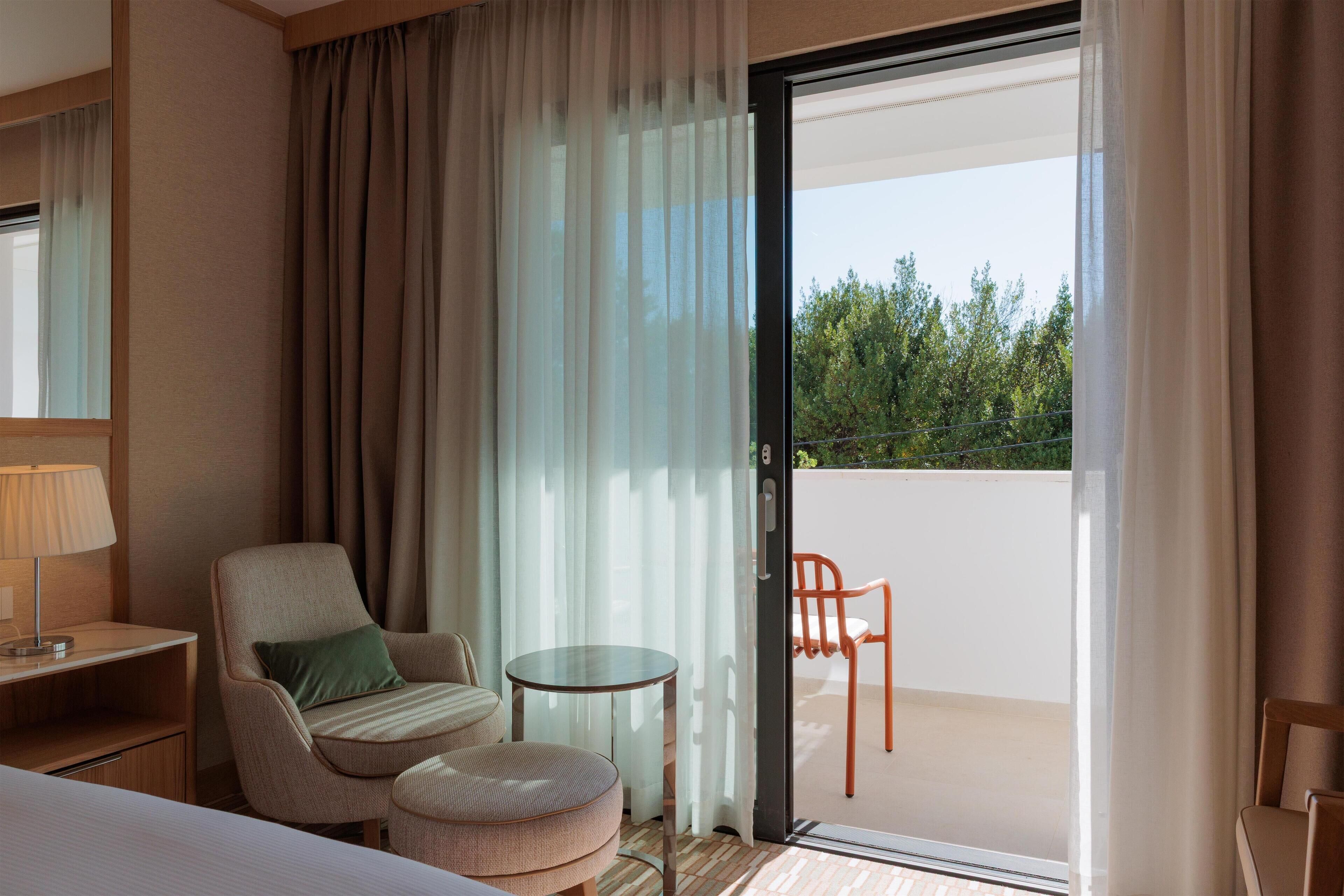 A hotel room with a bed, a chair, a small table, and a view of the outdoors through a sliding door.