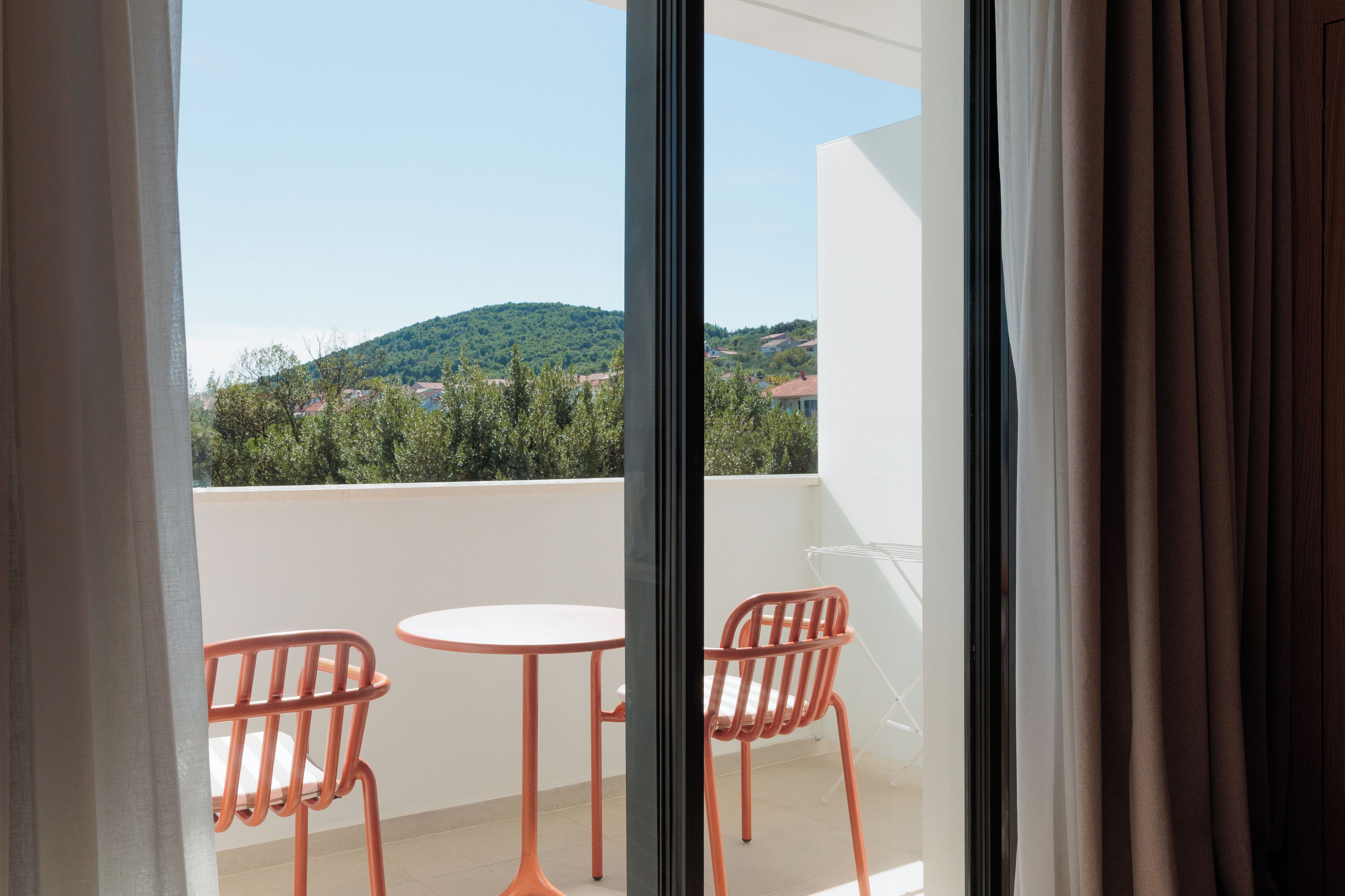 A balcony with two orange chairs and a small white table, overlooking a green hillside with buildings.