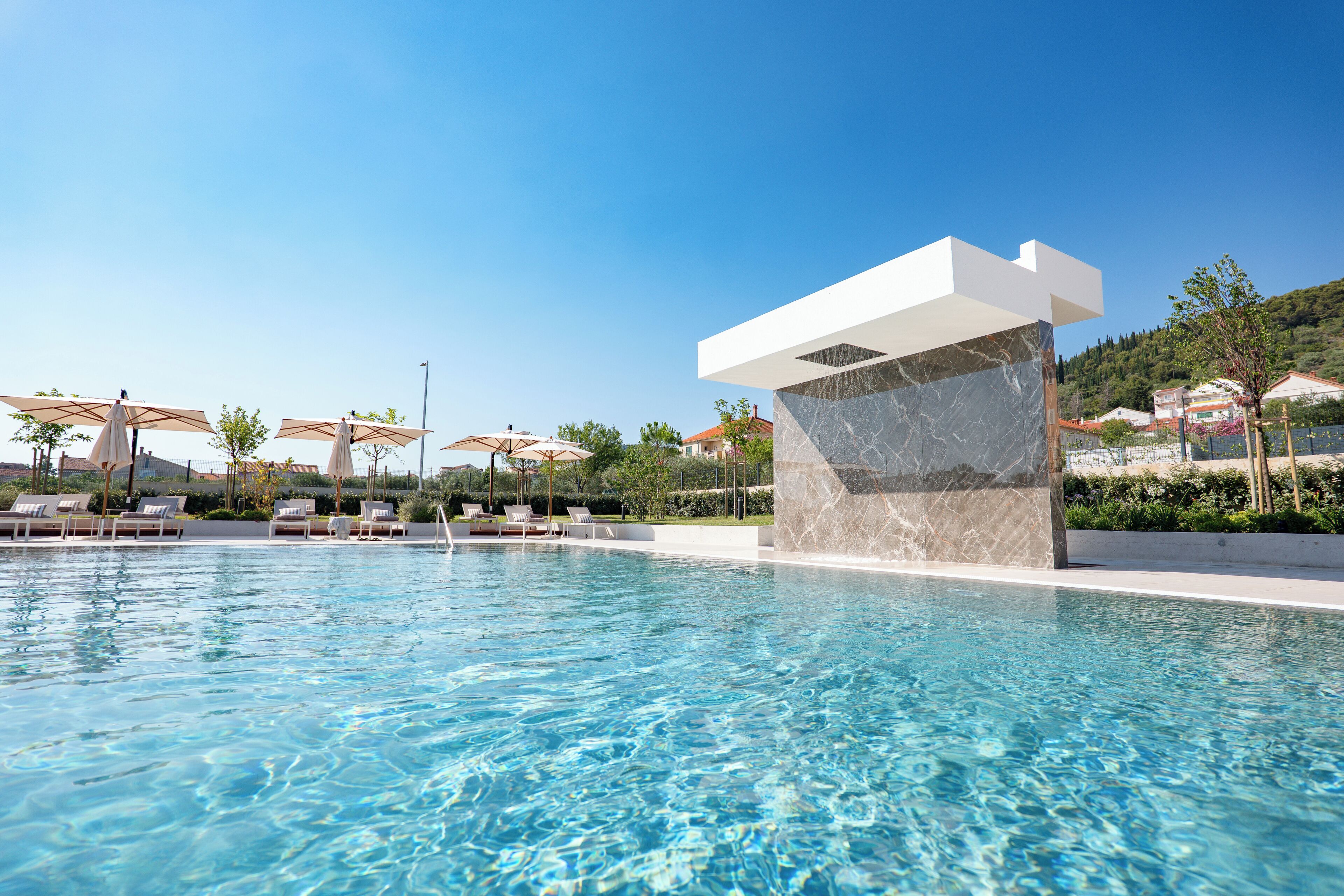 A clear swimming pool with lounge chairs and umbrellas, surrounded by a landscaped area.
