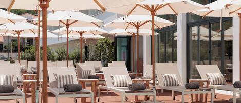 A sunlit outdoor lounge area with multiple white lounge chairs and beige umbrellas, arranged on a wooden deck.