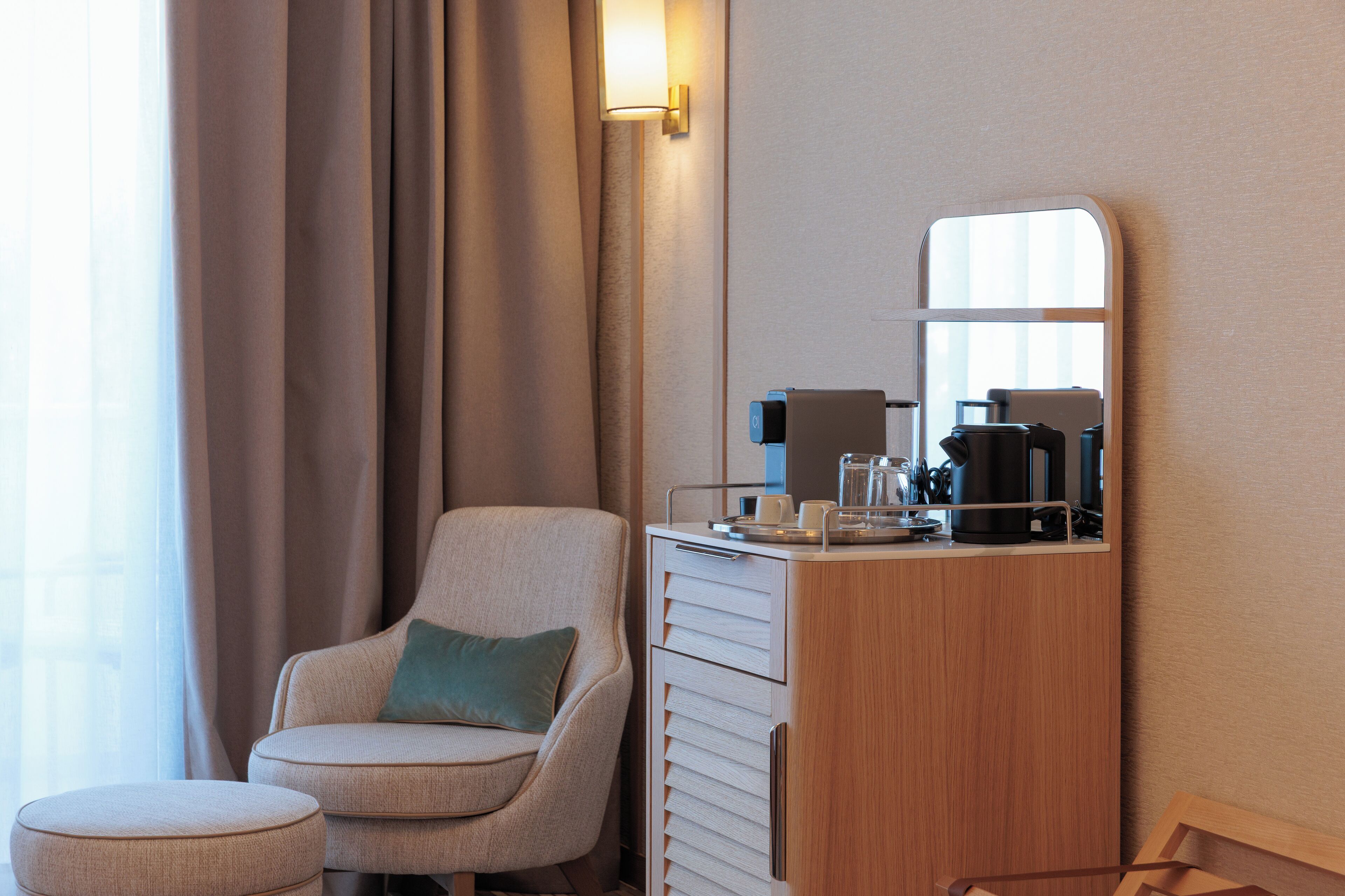 A hotel room with a sofa, a small ottoman, a wooden cabinet with a mirror, and a coffee maker.