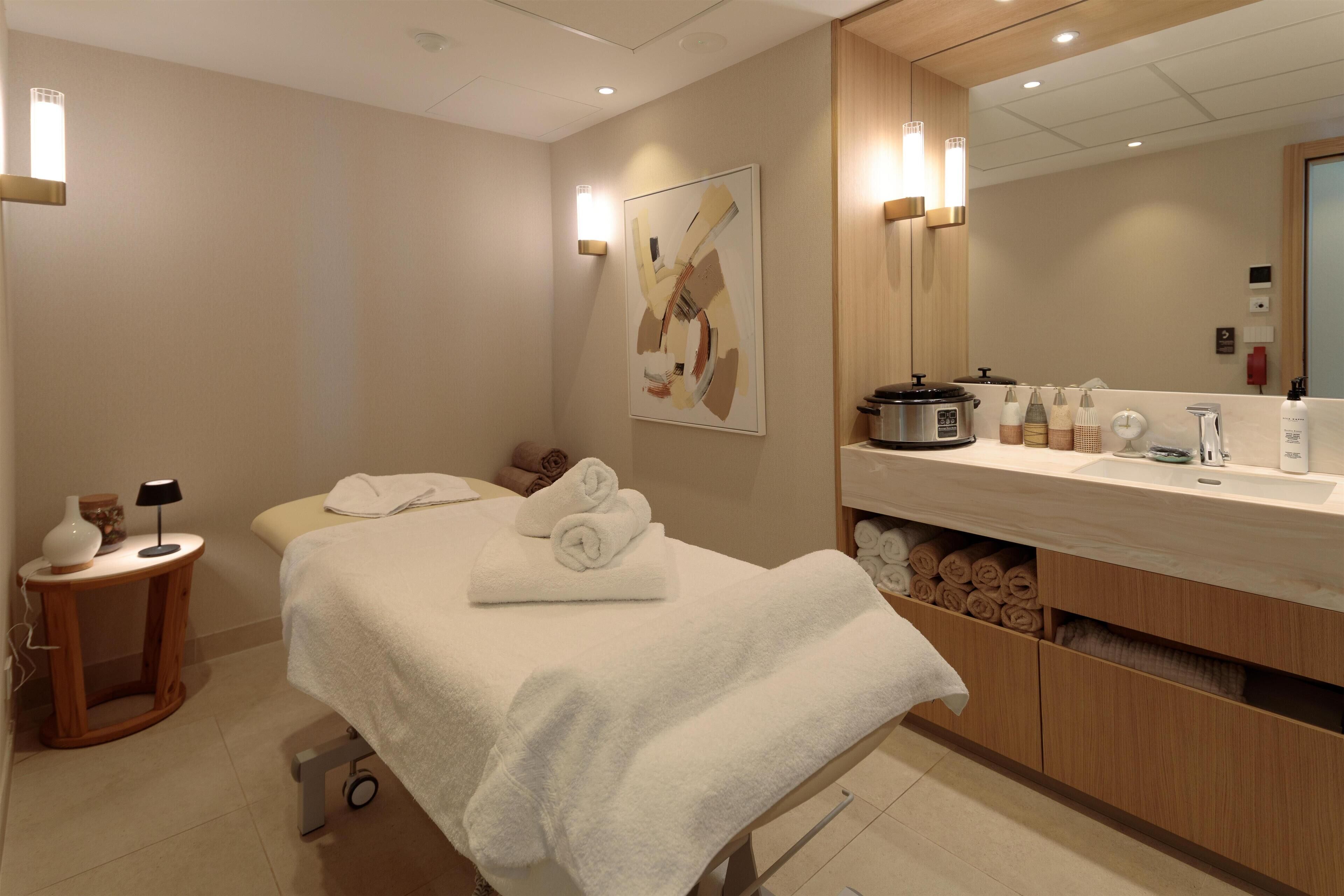 A modern hotel room with a bed, a bedside table with a lamp, a large mirror, and a counter with toiletries.