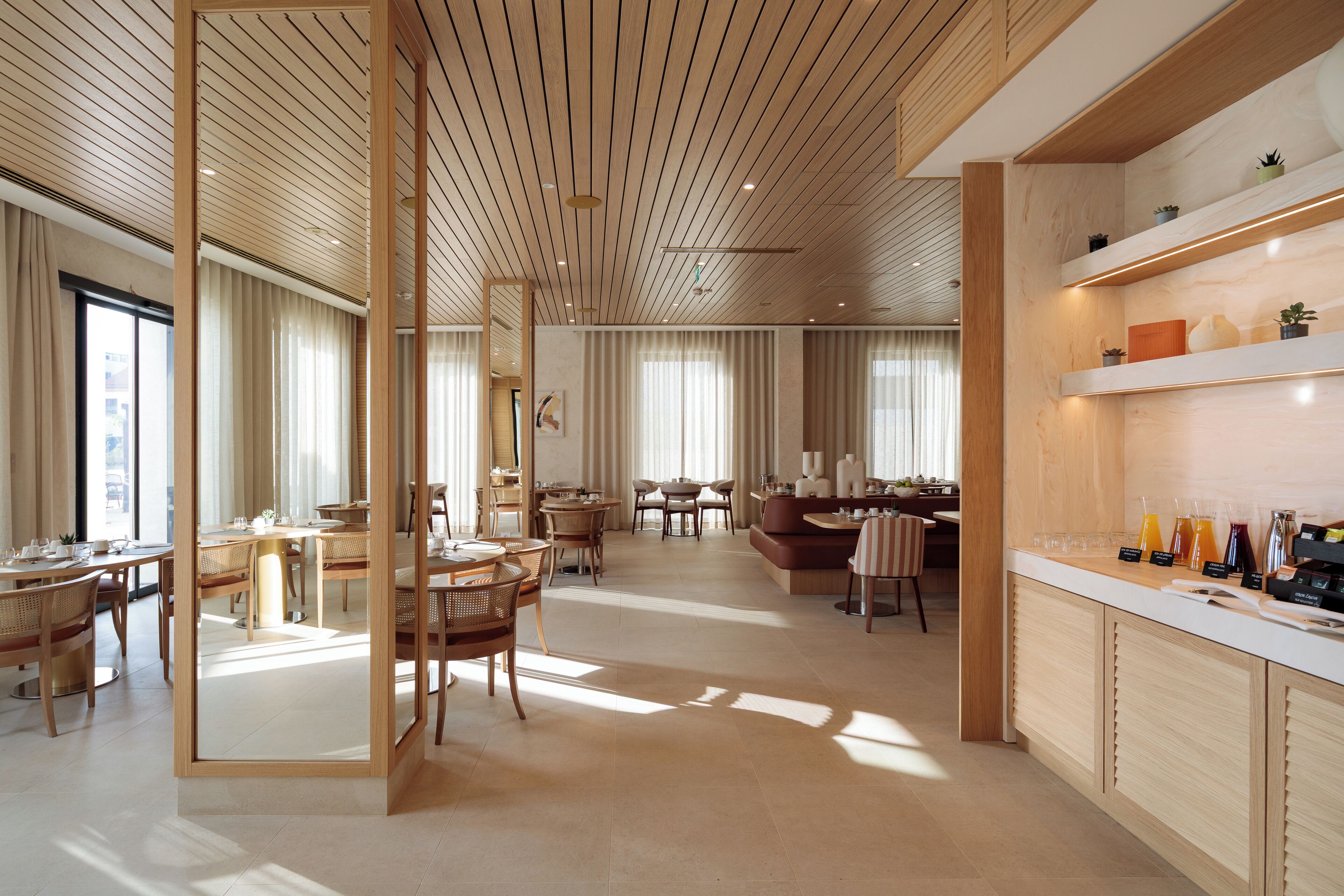 A spacious hotel lobby with wooden flooring, a central seating area, and a breakfast bar with various amenities.