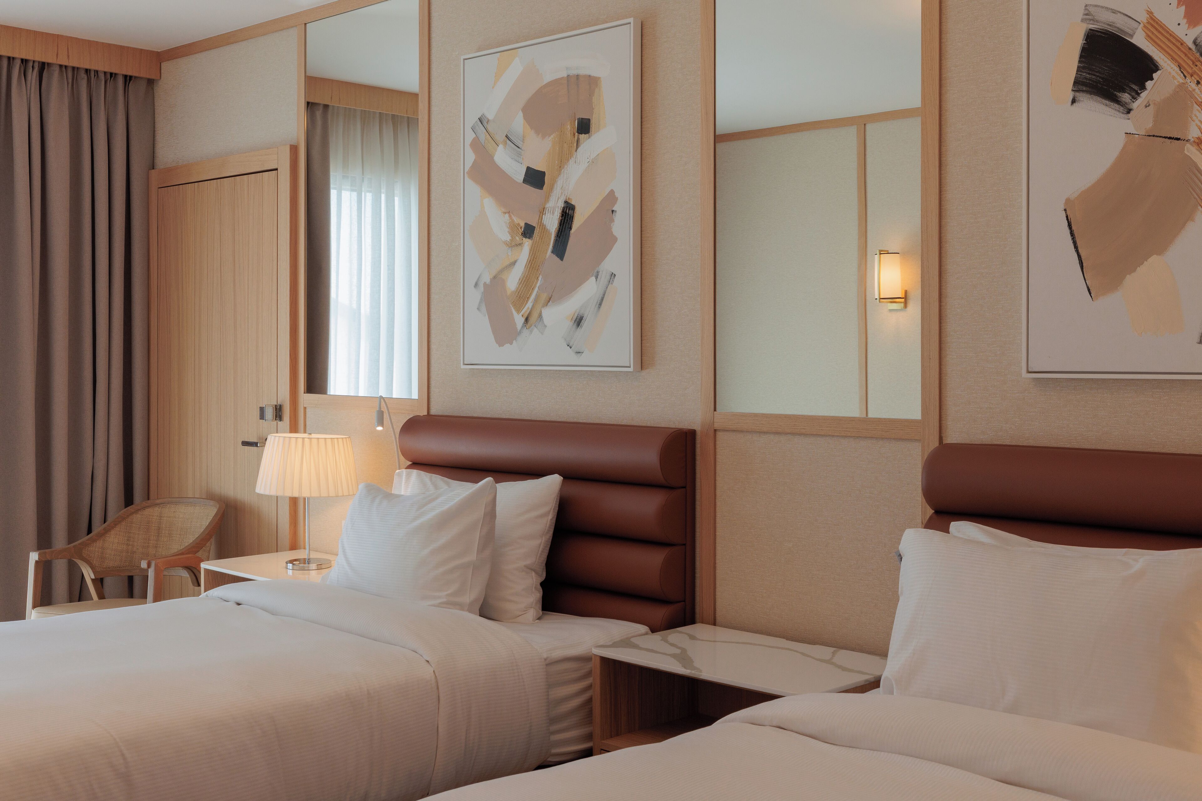 A hotel room with two beds, a chair, a lamp, and abstract art on the walls.