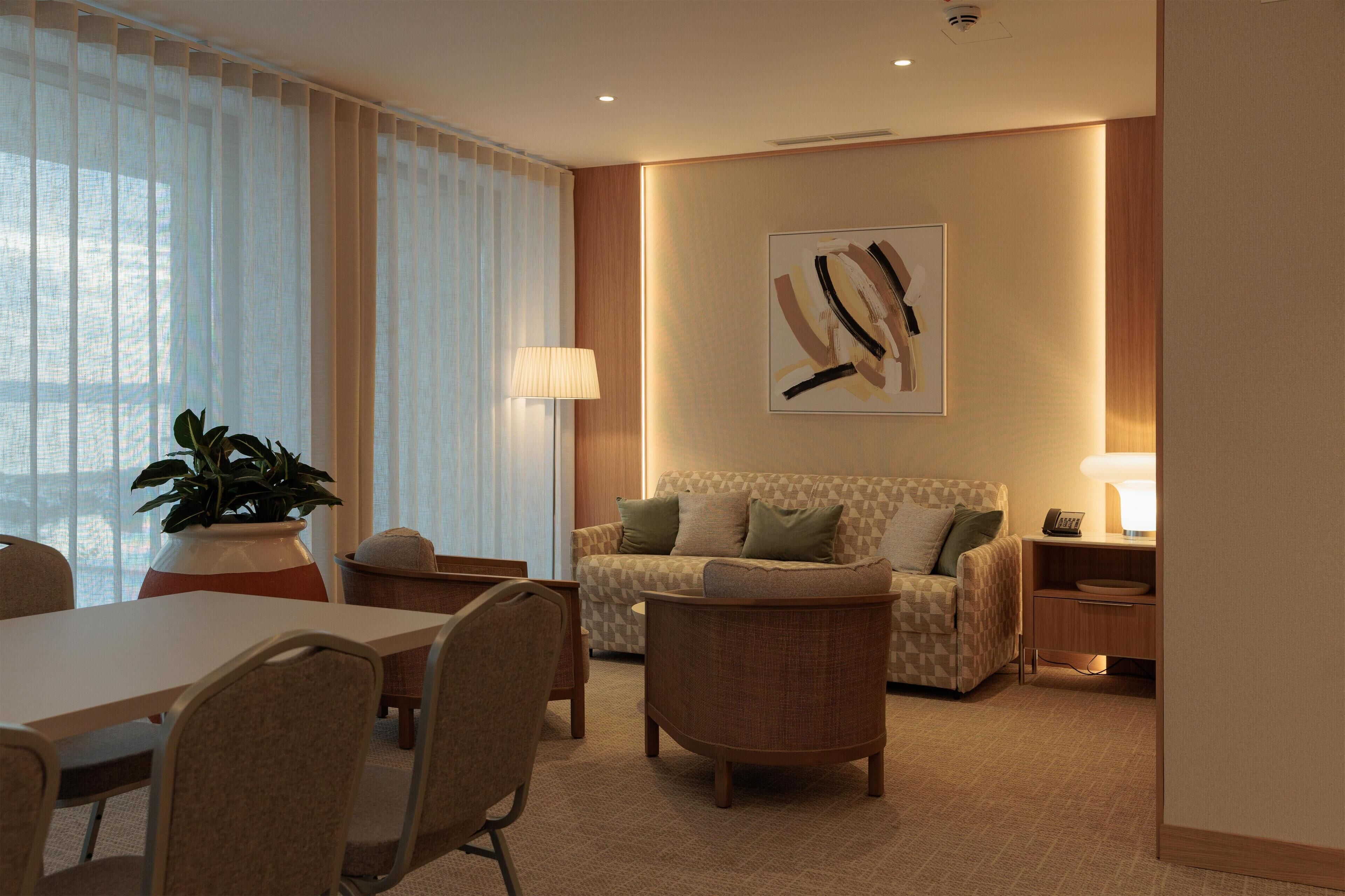 A modern hotel room with a dining area, a sofa set, a potted plant, and a painting on the wall.