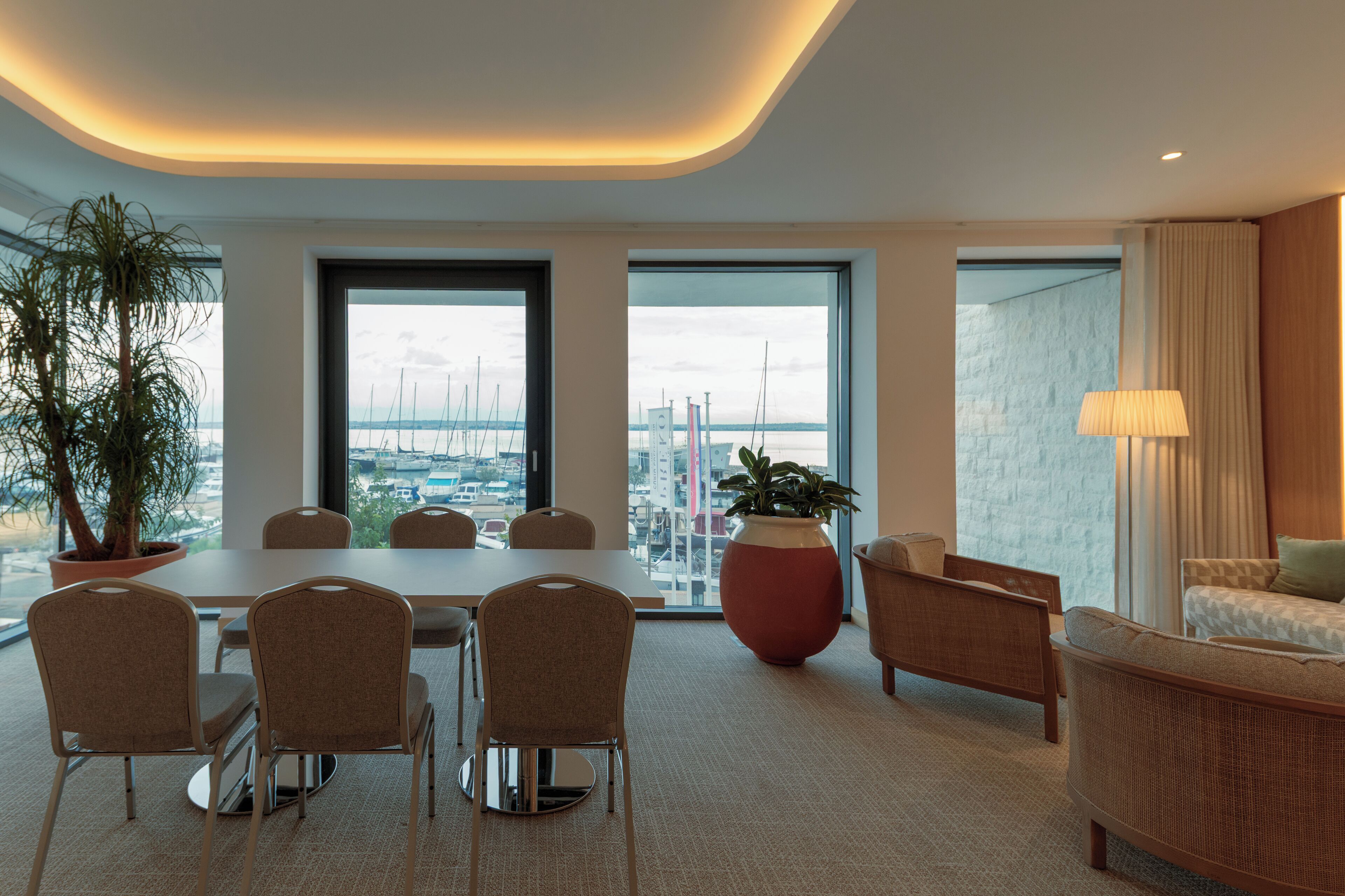 Une chambre d’hôtel moderne dotée d’une grande table à manger, de sièges confortables, d’une grande plante en pot et offrant une vue sur le paysage urbain grâce à de larges fenêtres.