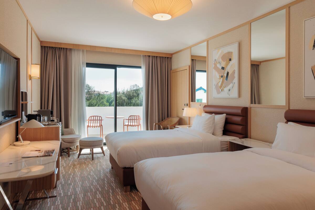 Une chambre d’hôtel avec deux lits, un bureau avec une chaise, une télévision et un balcon donnant sur un espace verdoyant.