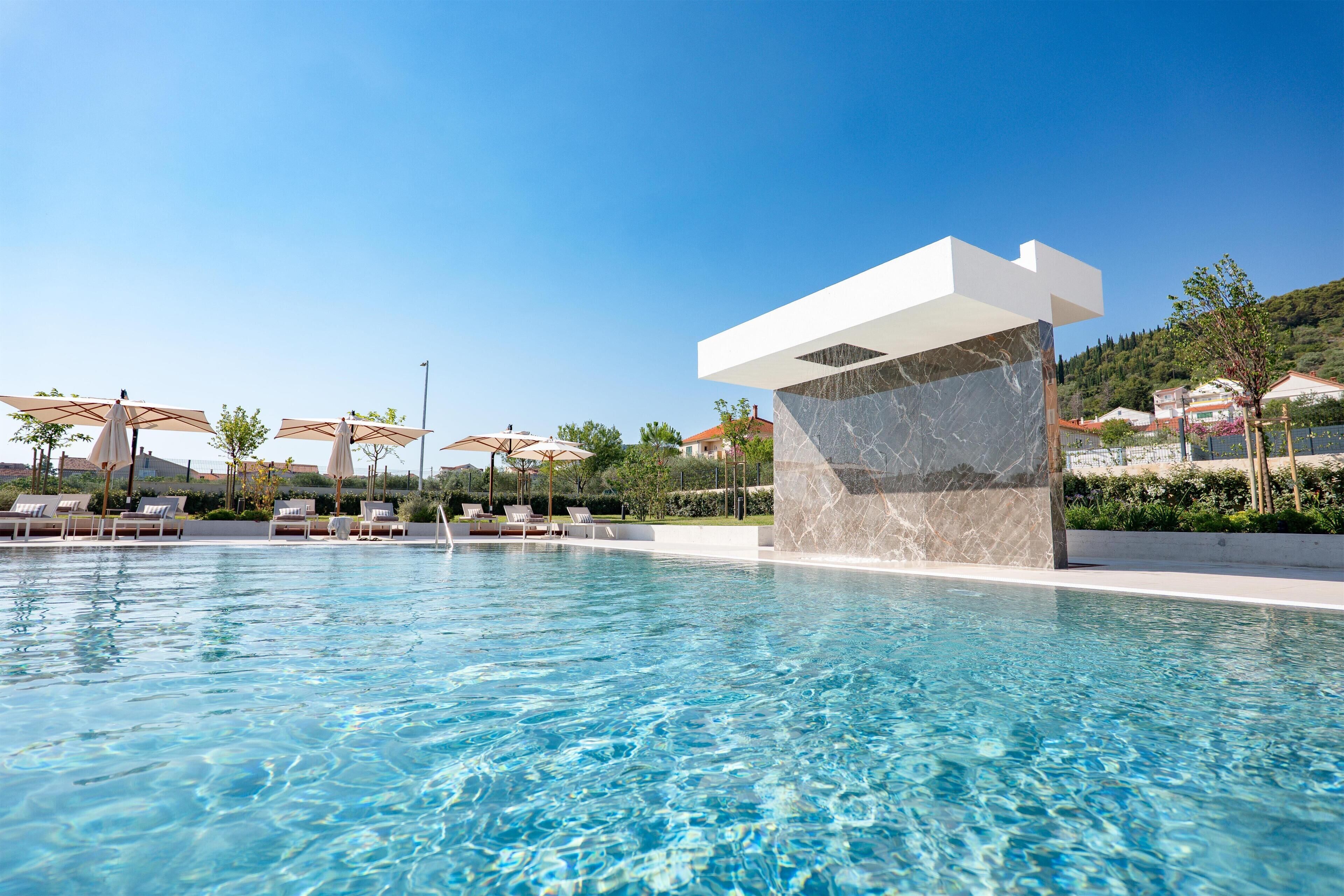 Piscina con sillas de descanso y sombrillas, rodeada de un área ajardinada con árboles y cielo despejado.