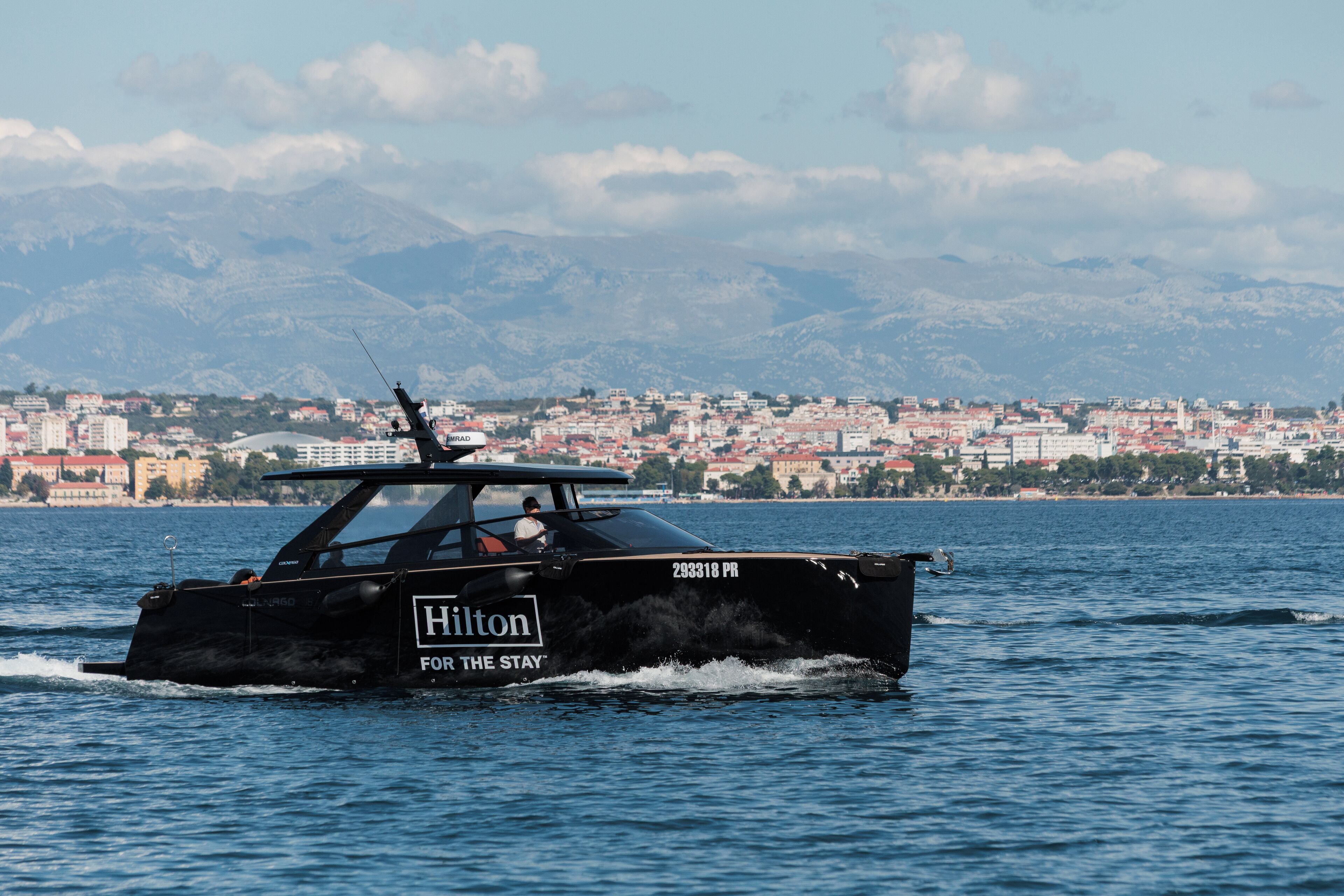 Ein schwarzes Boot mit der Aufschrift "Hilton For The Stay" fährt auf dem Wasser, im Hintergrund sind Stadt und Berge zu sehen.