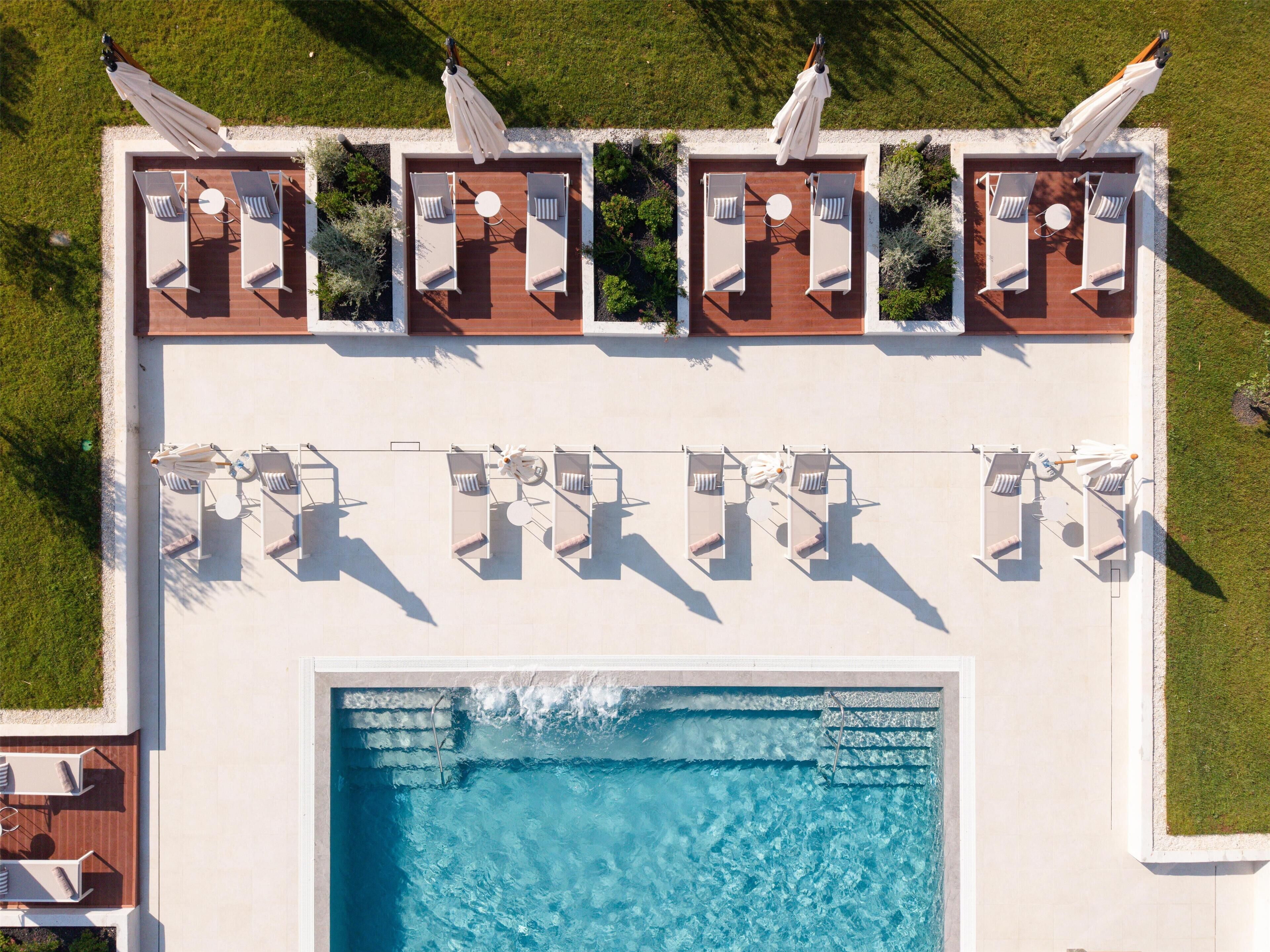 Vista aérea de una zona de piscina con múltiples sillas de descanso y sombrillas, rodeada por un solárium blanco.